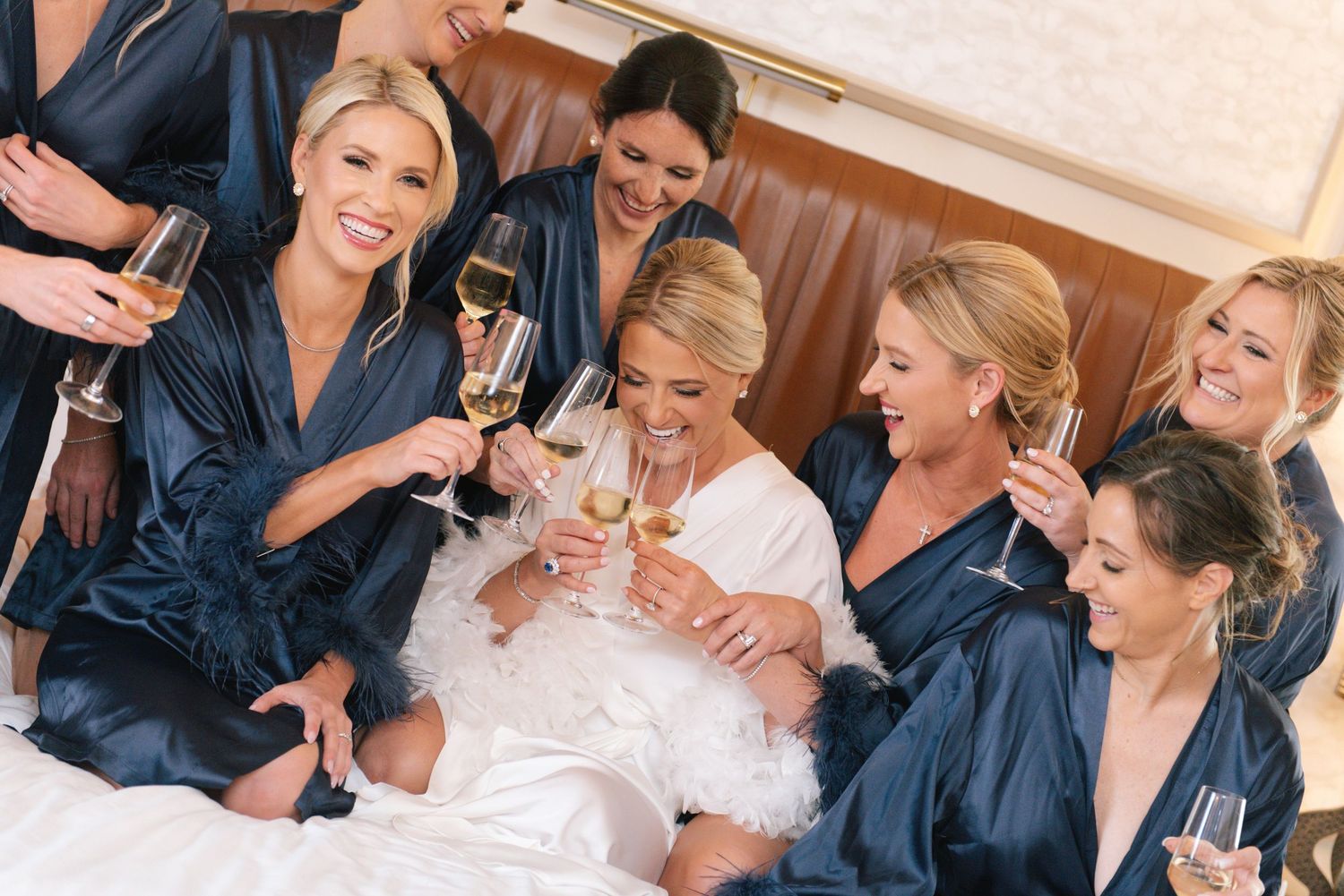 Bridesmaids in navy satin robes toasting with champagne glasses alongside the bride on her wedding morning.