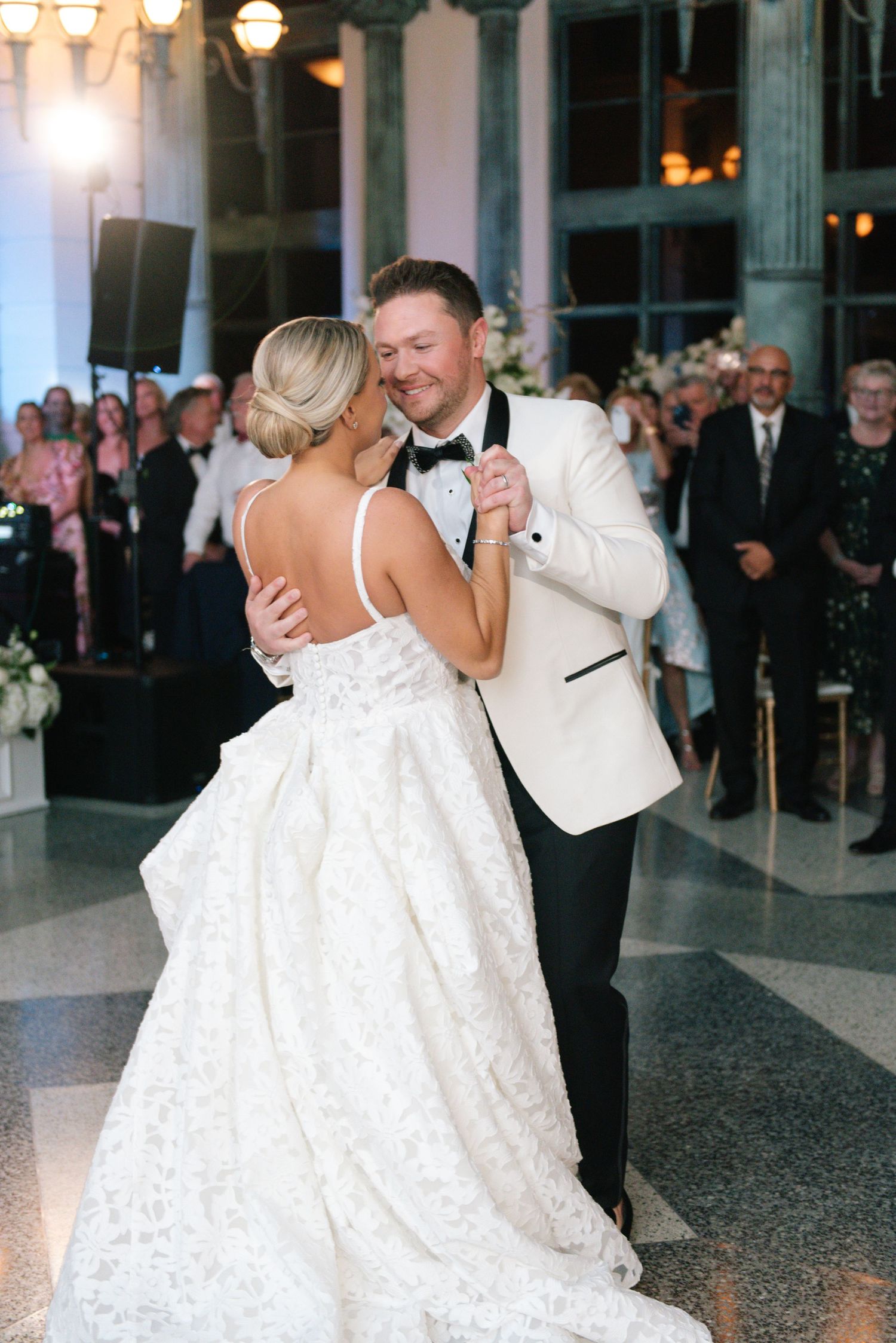 Bride and groom share their first dance, groom in white tuxedo jacket, bride in backless lace wedding gown.