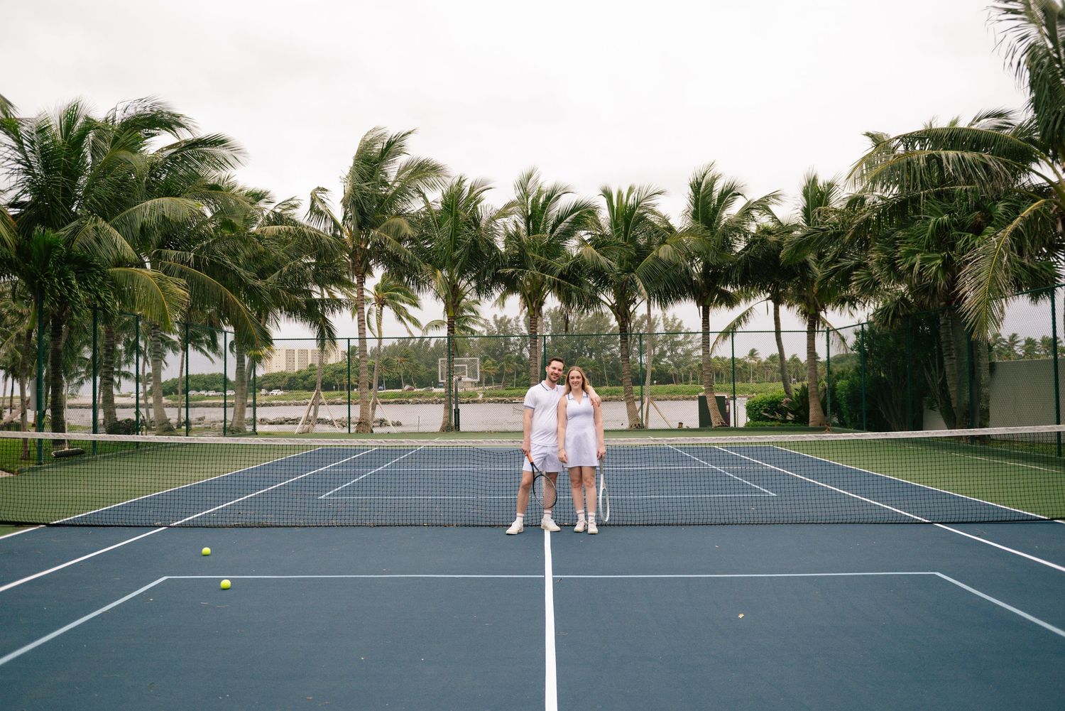 Empty tennis court surrounded by palm trees under cloudy skies.