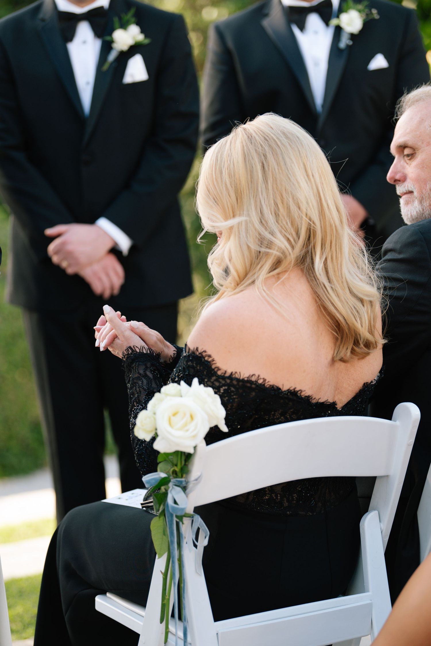 Wedding party members in black tuxedos and formal attire stand together during an outdoor ceremony.