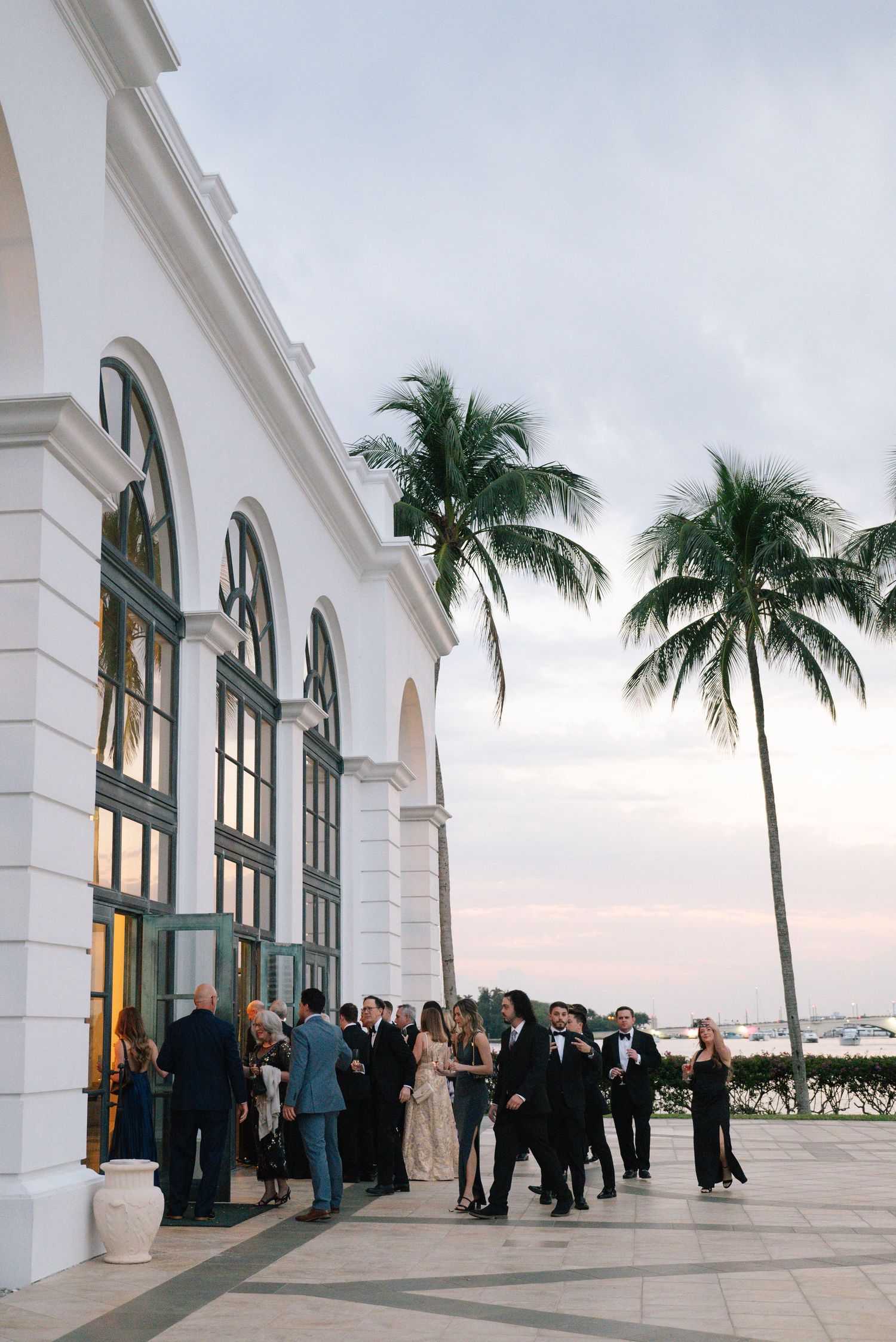 Elegant white building with arched windows and palm trees against cloudy sky during outdoor wedding reception.