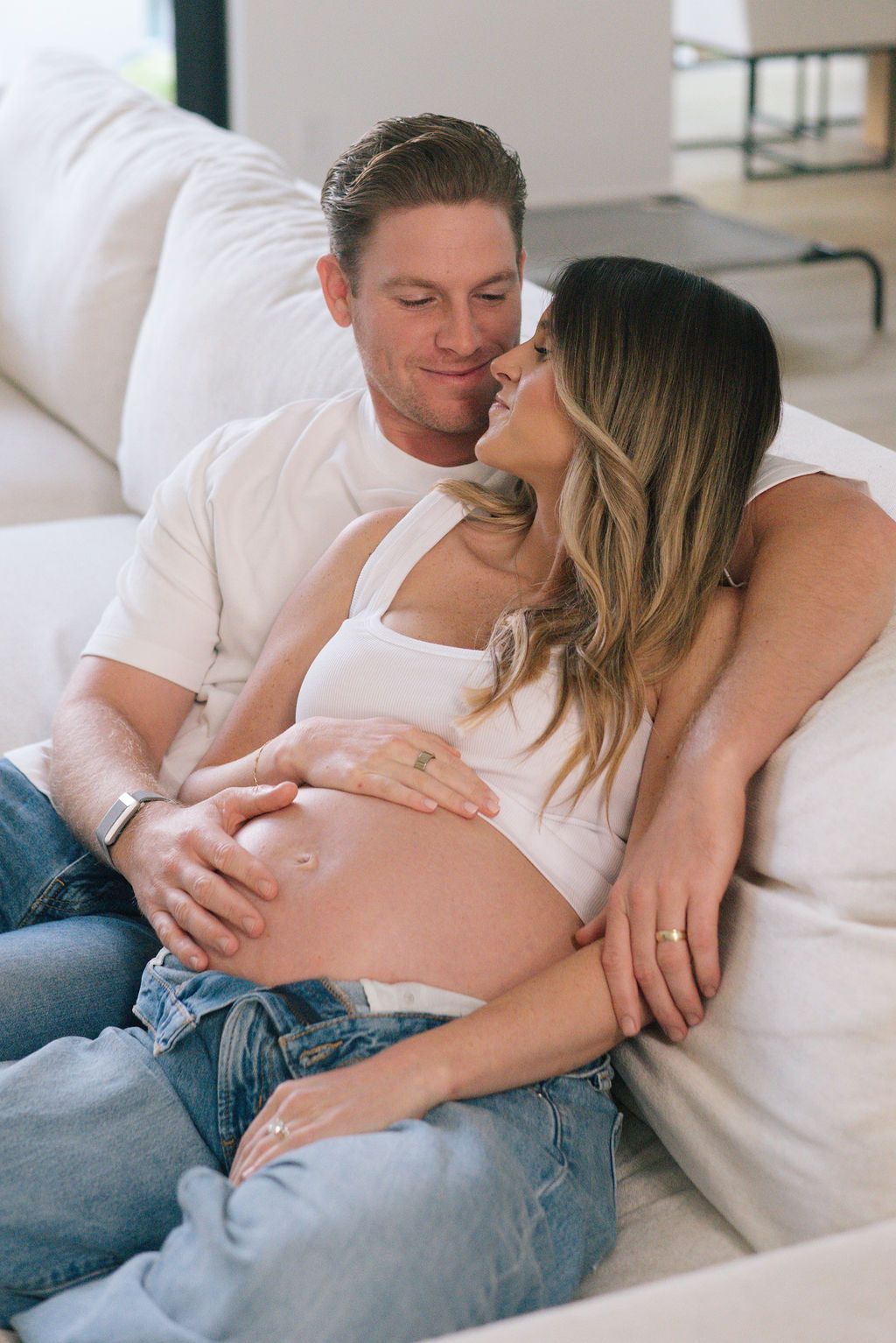 Couple sharing an intimate moment on a white couch while looking at ultrasound photos.