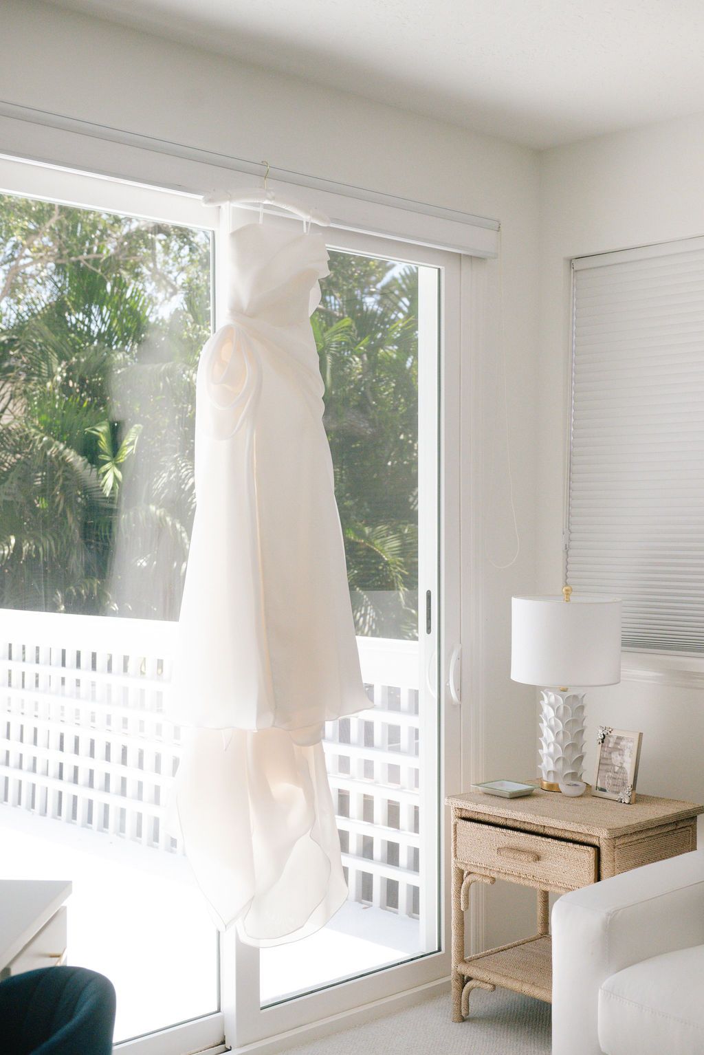 White wedding dress hanging by bright window with palm trees visible outside.