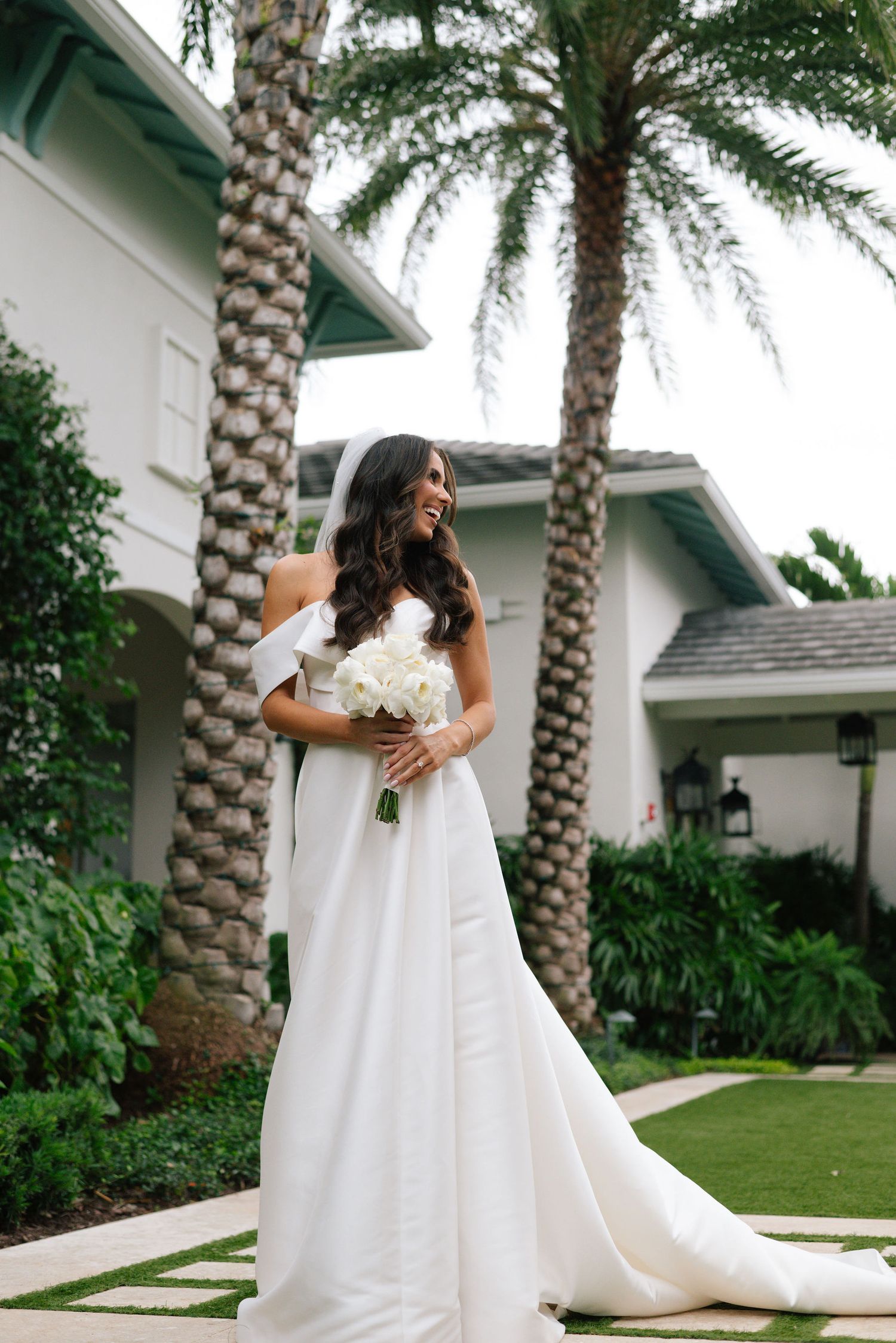 A flowing white wedding gown with a dramatic train is showcased against a tropical palm-lined garden setting.
