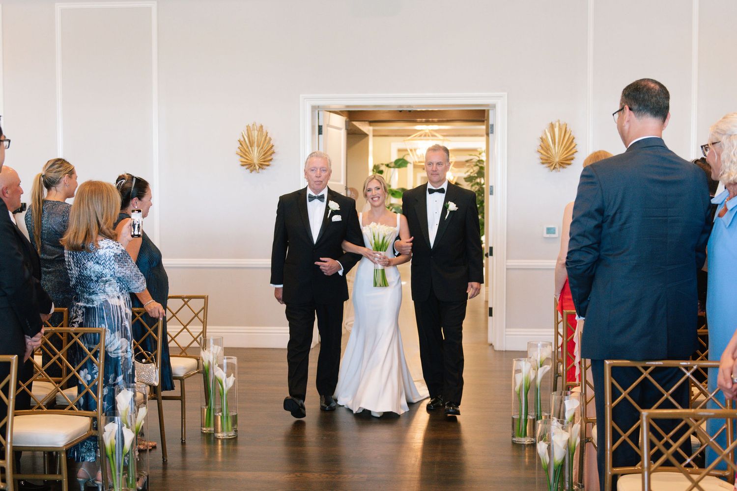 Elegant wedding ceremony processional in a bright indoor venue with white walls and gold sconces.