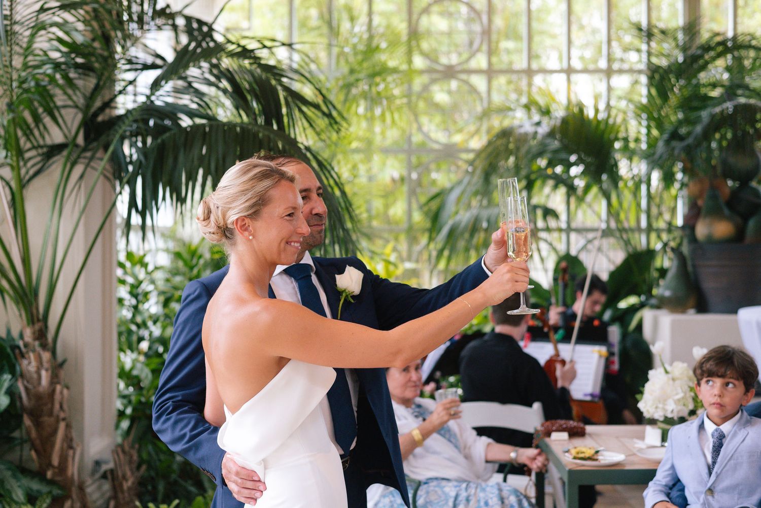 Wedding celebration in a sunlit conservatory with guests raising champagne glasses.