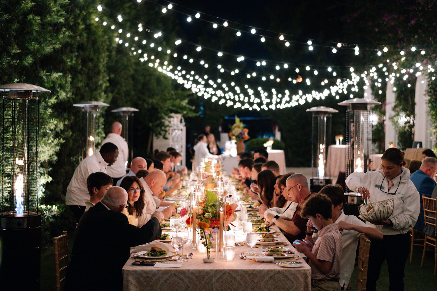 Long dinner table set under romantic string lights in an outdoor garden setting.