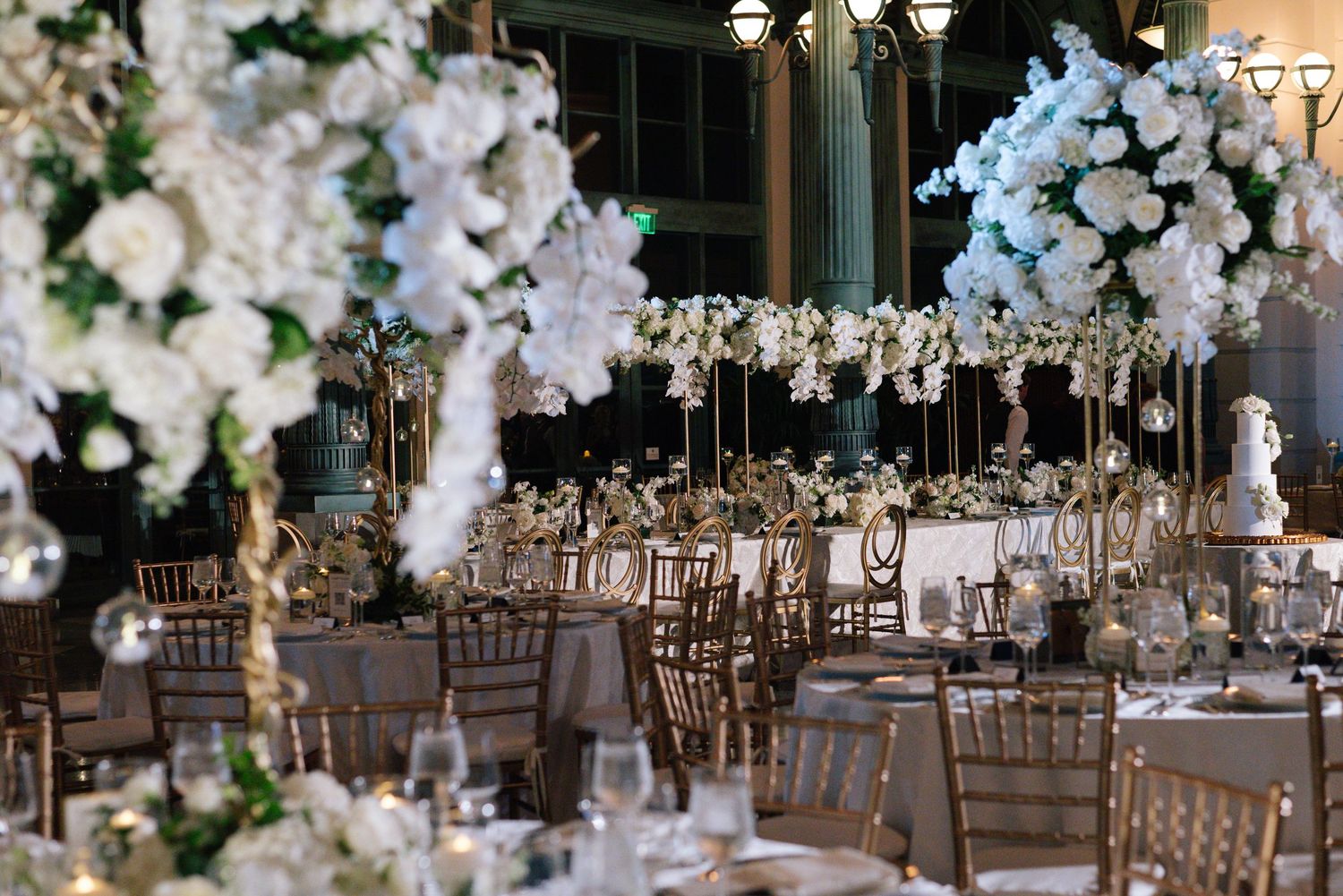 Luxurious wedding reception hall with towering white floral arrangements and gold chiavari chairs under ornate ceiling.