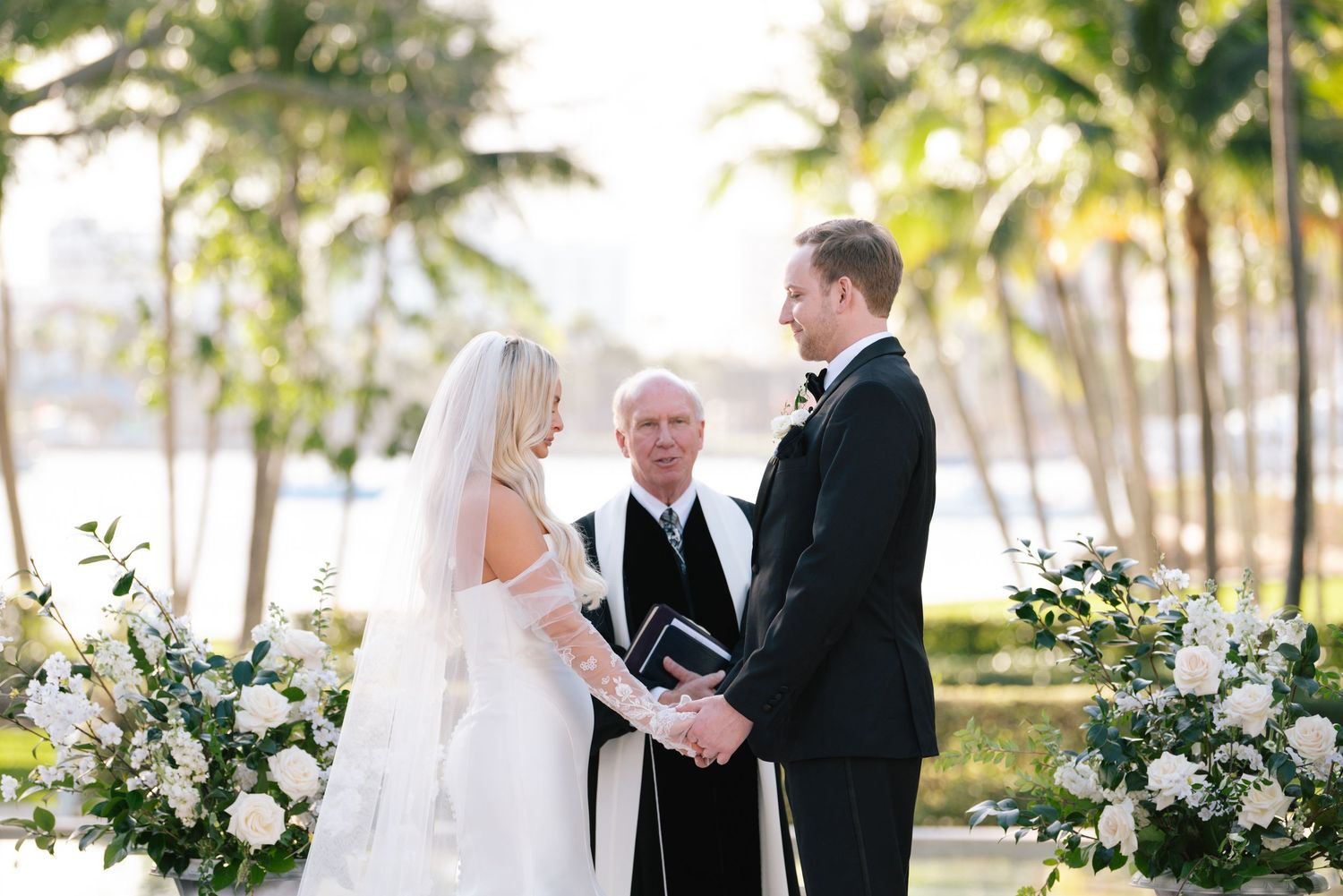 Wedding ceremony taking place outdoors with palm trees and white floral arrangements creating a tropical setting.