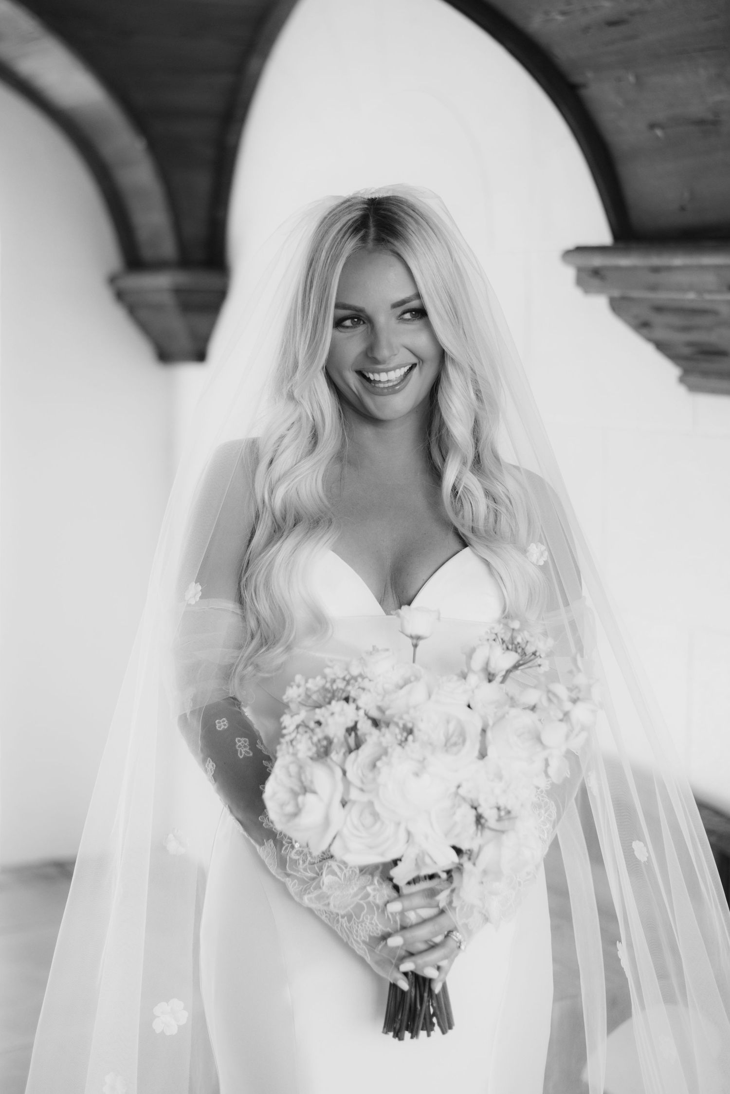 Black and white portrait of a bride in a fitted white gown holding a bouquet of flowers under an arched doorway.