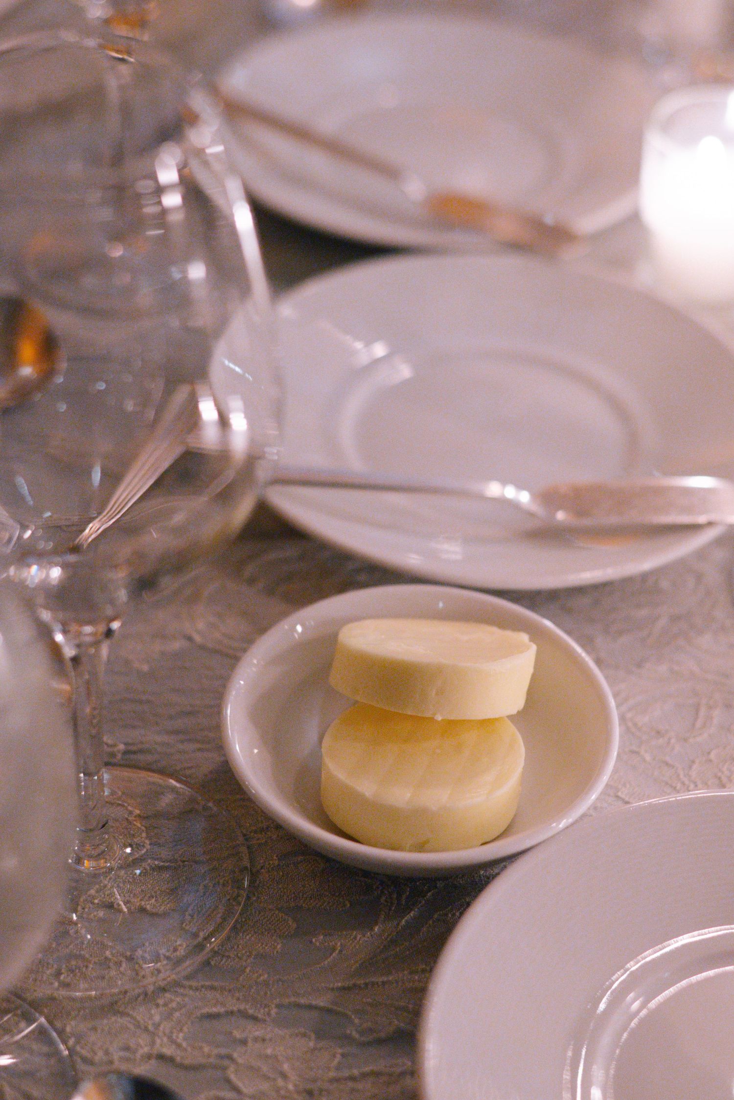 Place setting with white plates, wine glasses and butter pats on an ornate tablecloth.