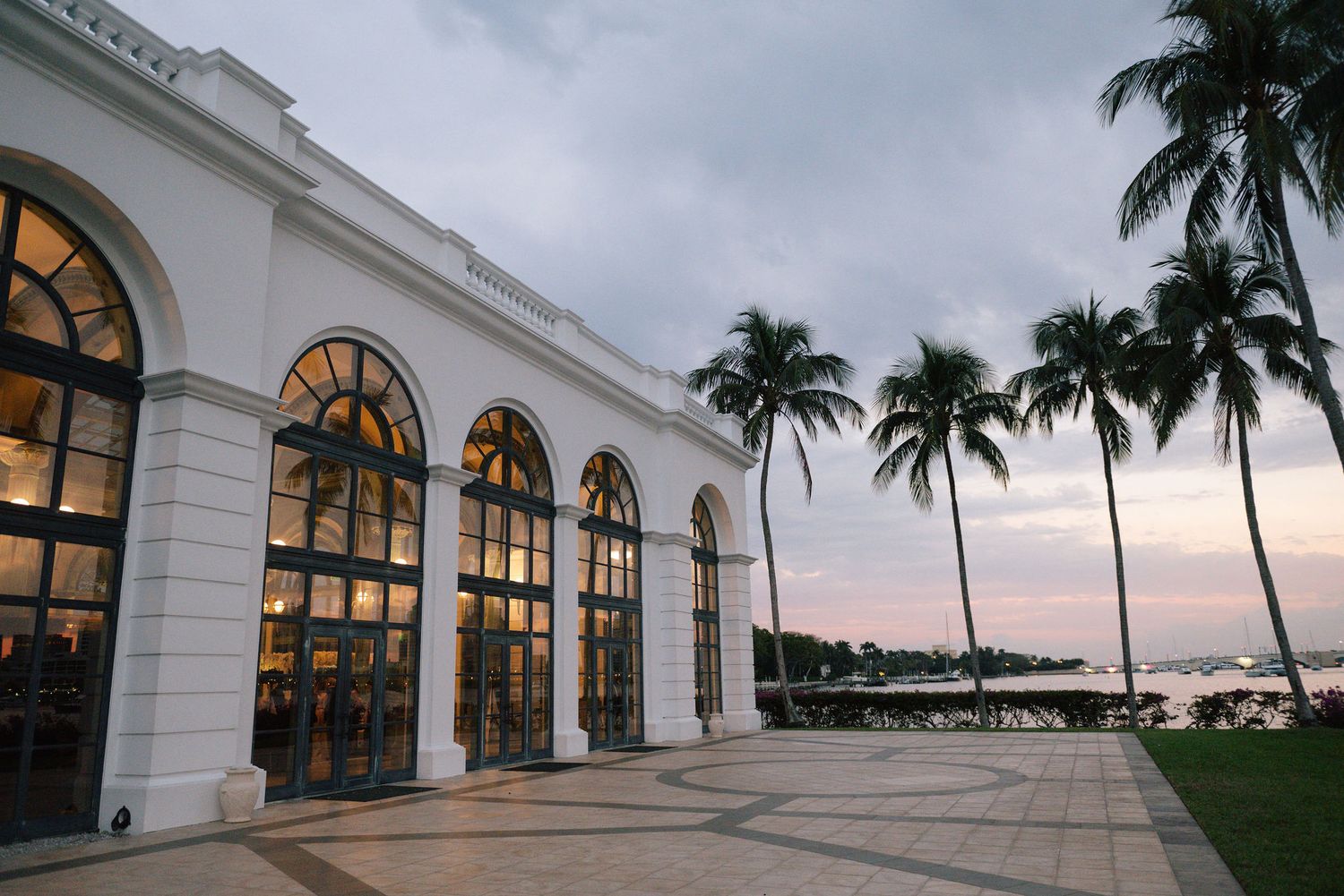 Exterior view of white colonial building with arched windows and palm trees at sunset.