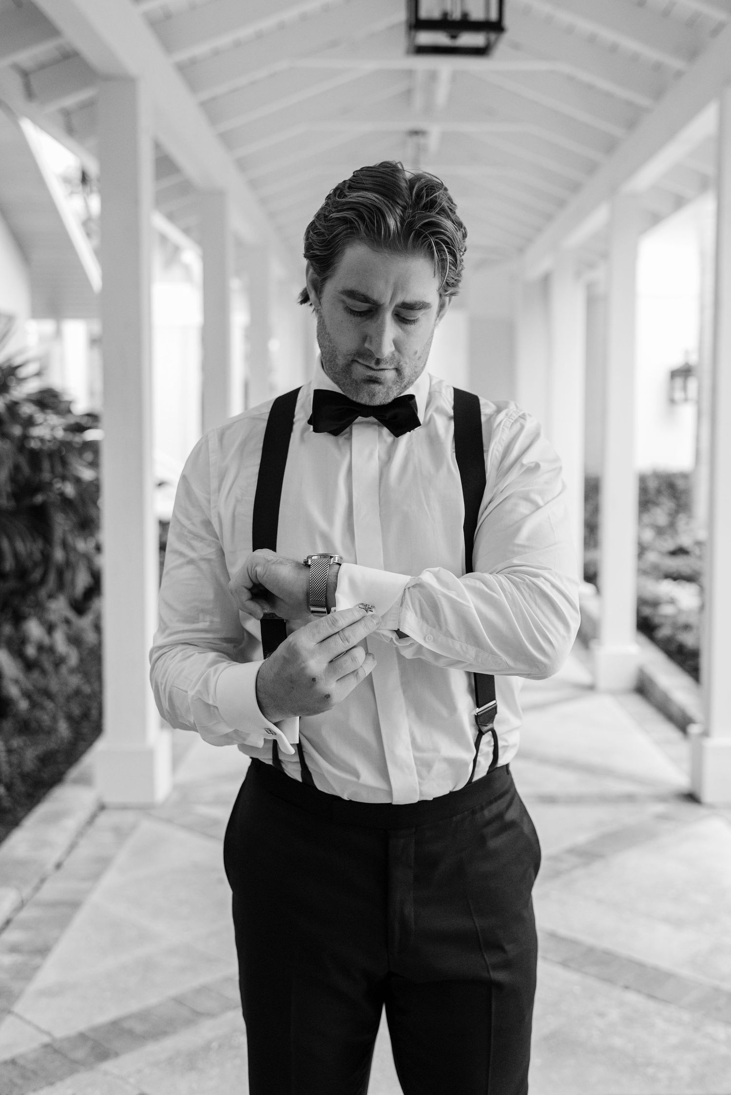 Groom in classic white shirt and suspenders checks time on watch in covered walkway.