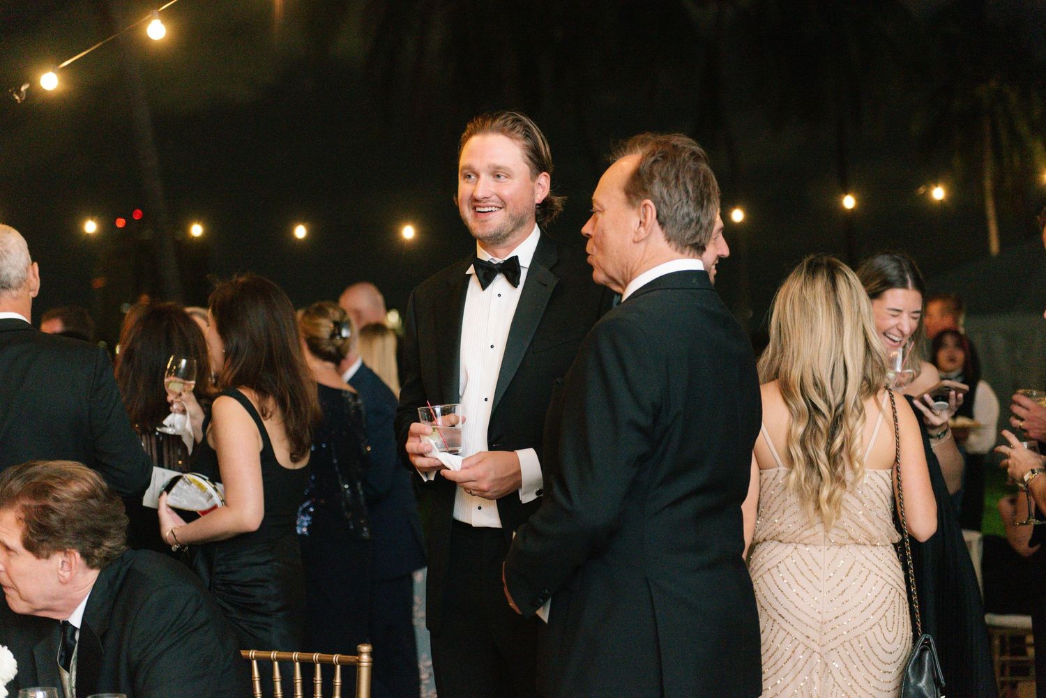 Guests in formal black tie attire socializing at elegant outdoor wedding reception.