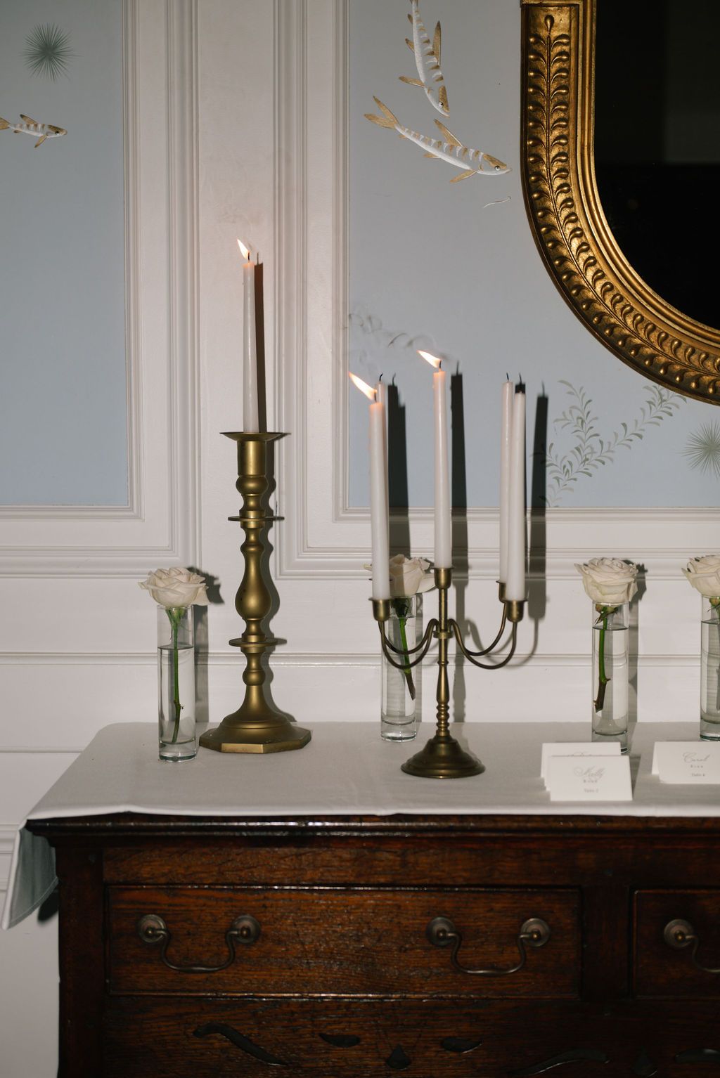 Vintage wooden sideboard with brass candlesticks and ornate gold mirror in a formal setting.