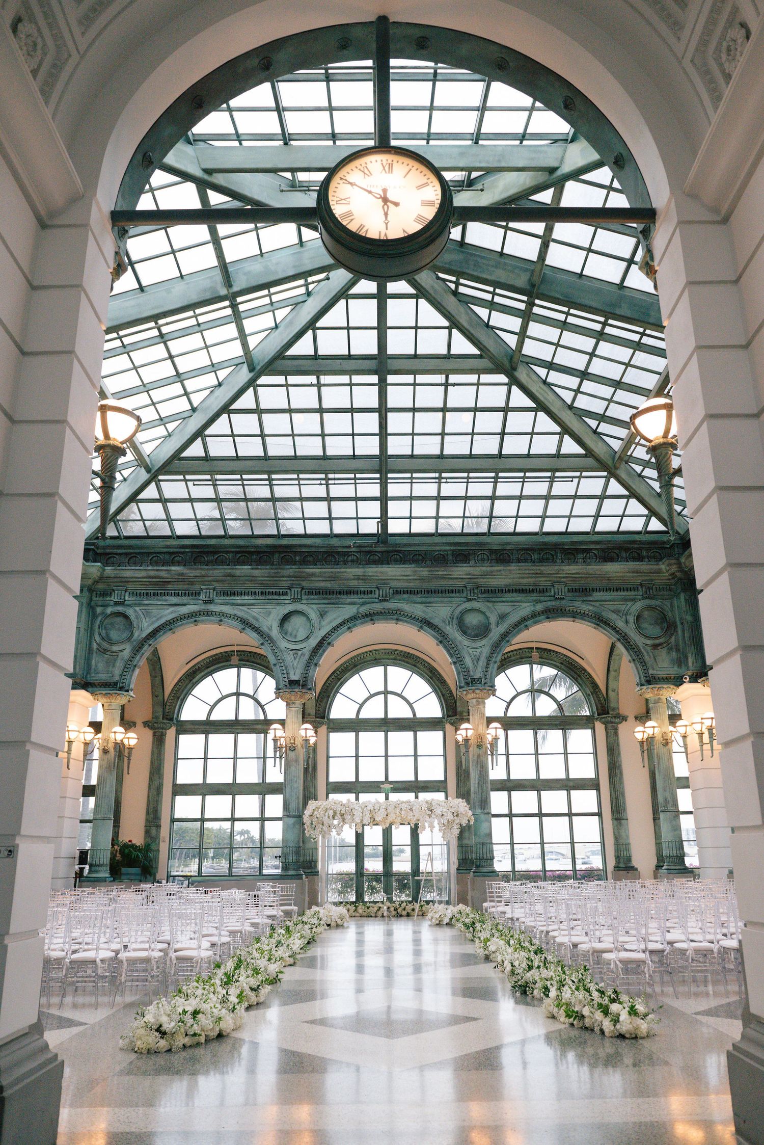 Luxurious wedding venue with glass ceiling, arched windows and white chairs set up for ceremony.