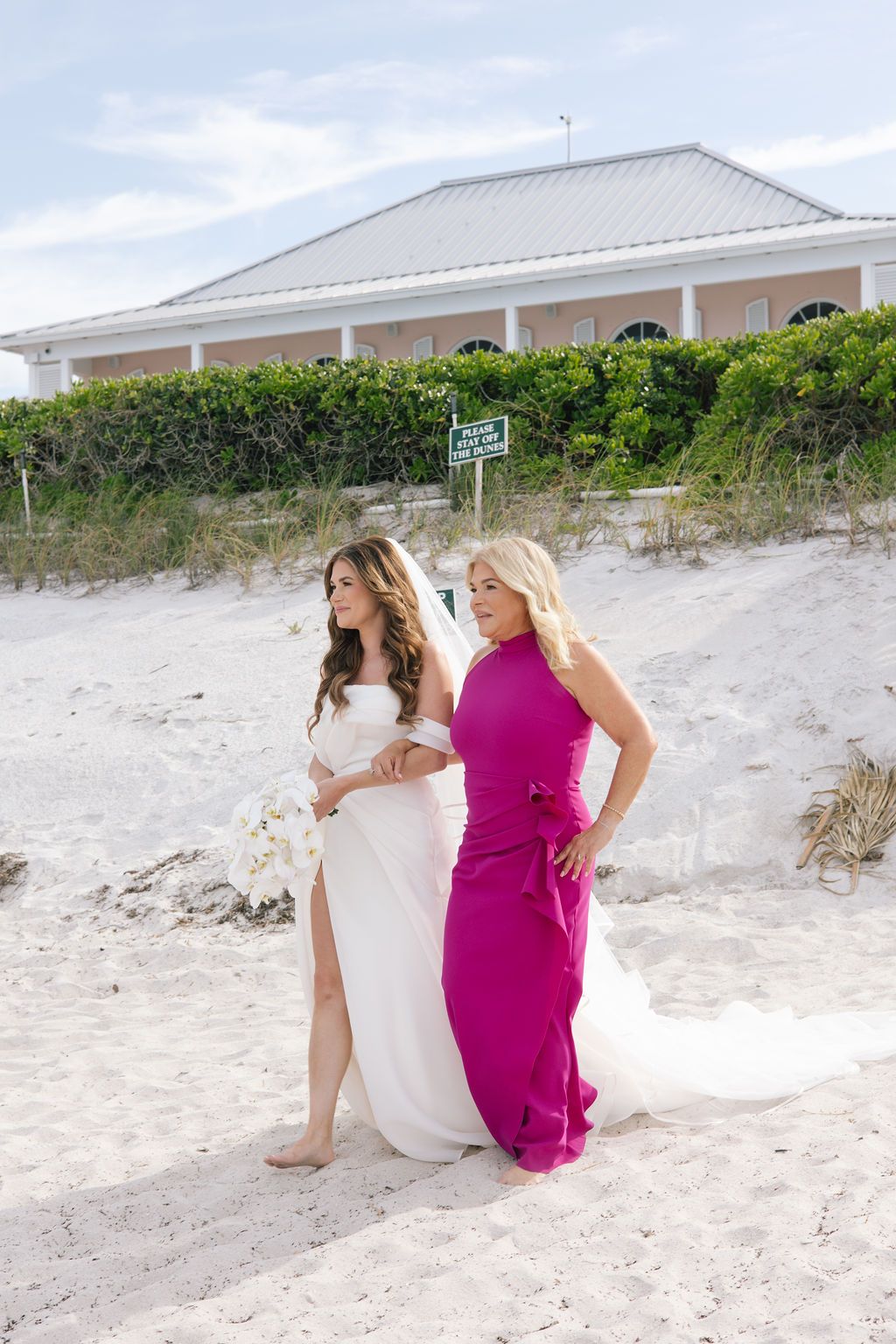 Wedding party members in pink dresses sitting on sandy dunes near beach houses.