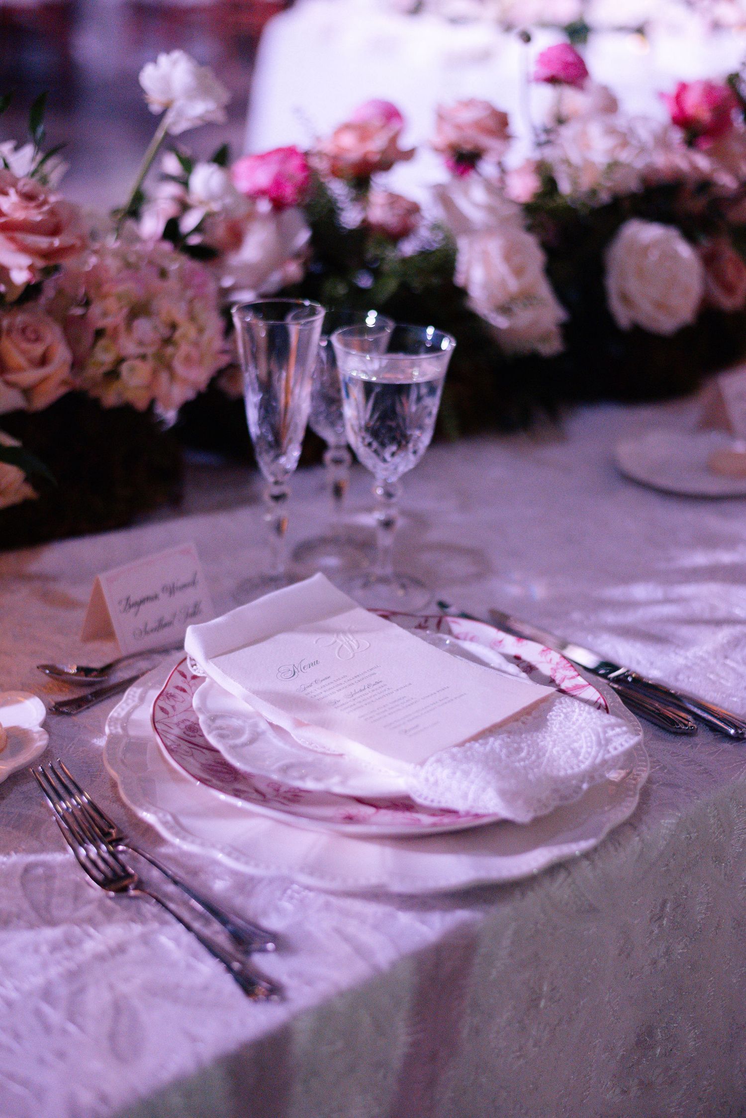 Close-up of formal place settings with pink floral arrangements and white tablecloths at a wedding reception.