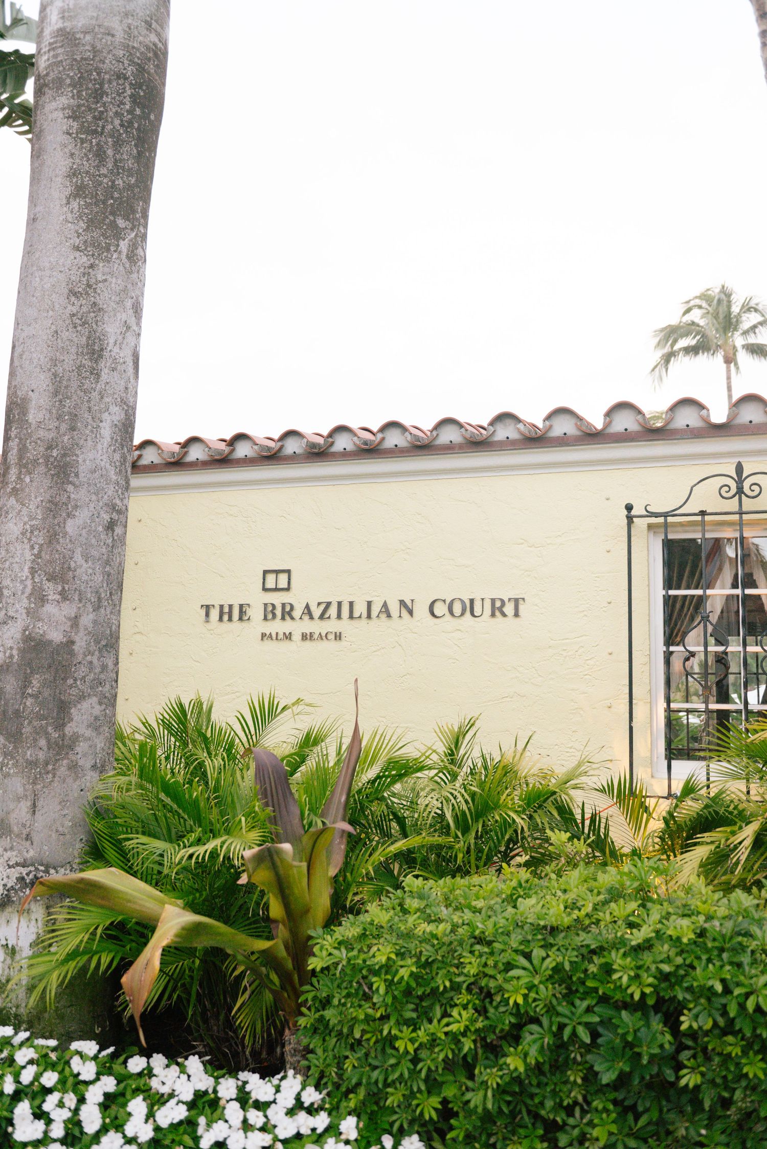 A sign for 'The Brazilian Court' with palm trees and lush tropical landscaping in front of a cream-colored building.