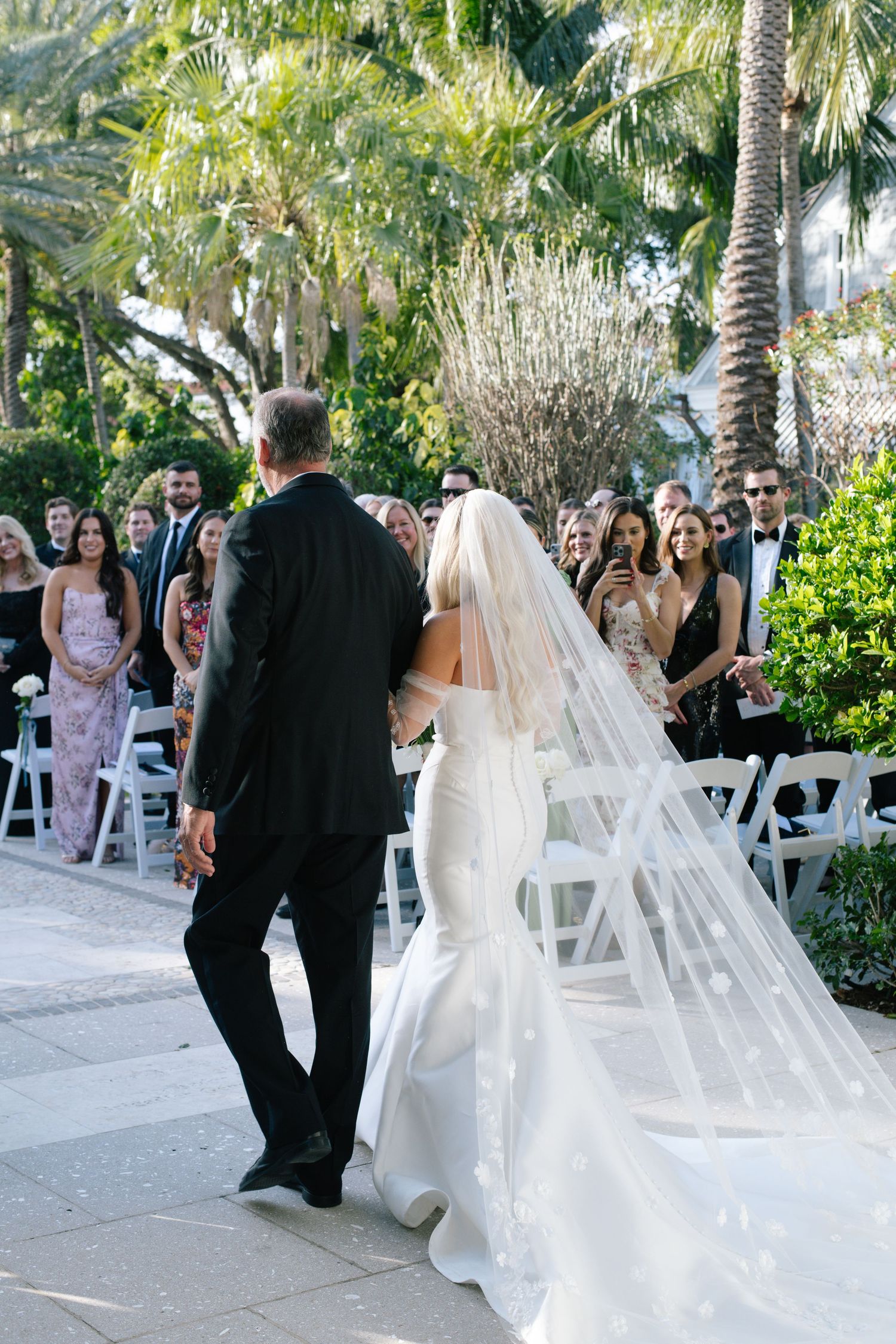 Wedding ceremony guests watch as a couple proceeds down the aisle lined with white chairs and tropical landscaping.