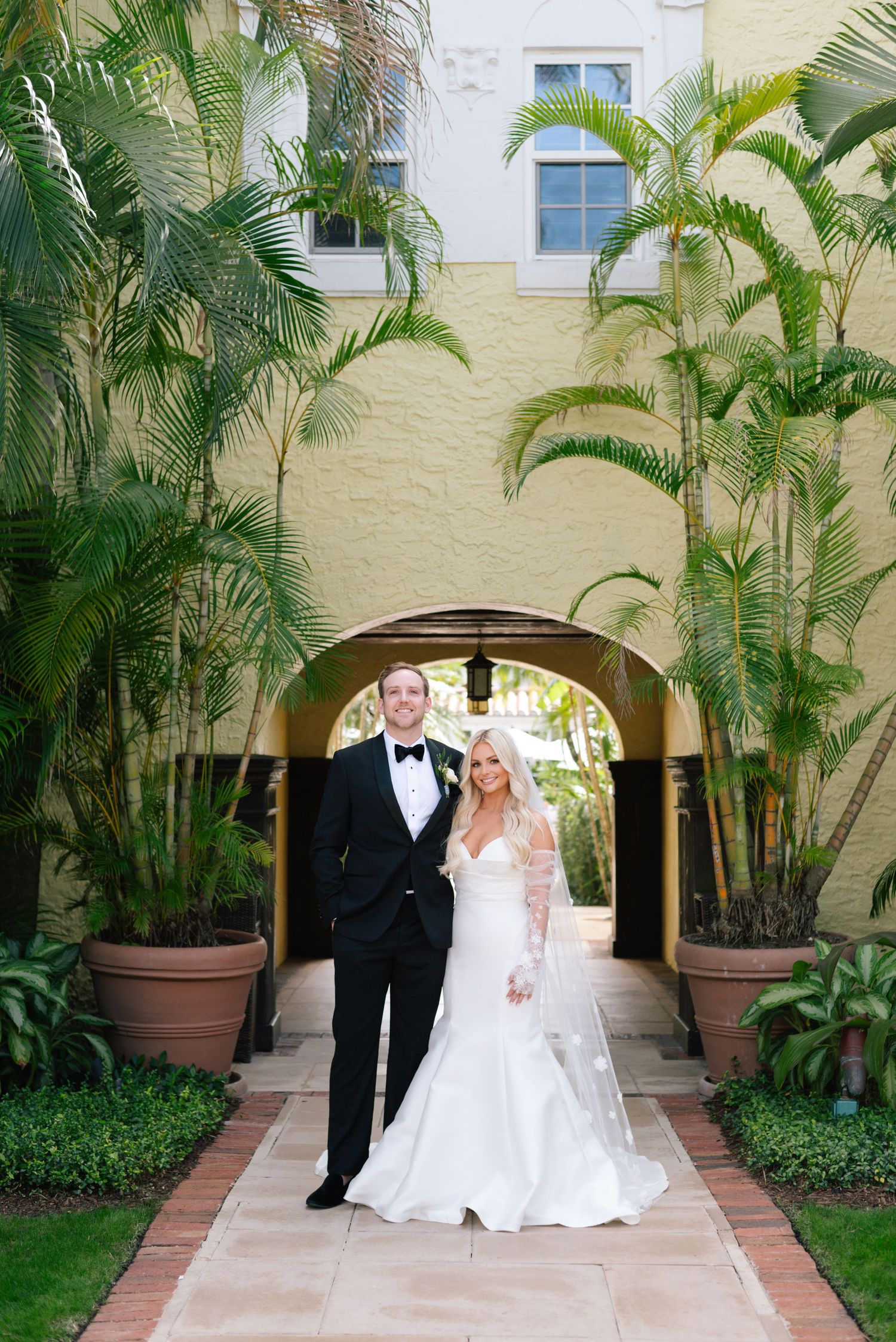 Elegant wedding couple poses in a lush tropical courtyard with tall palm trees and arched walkways.