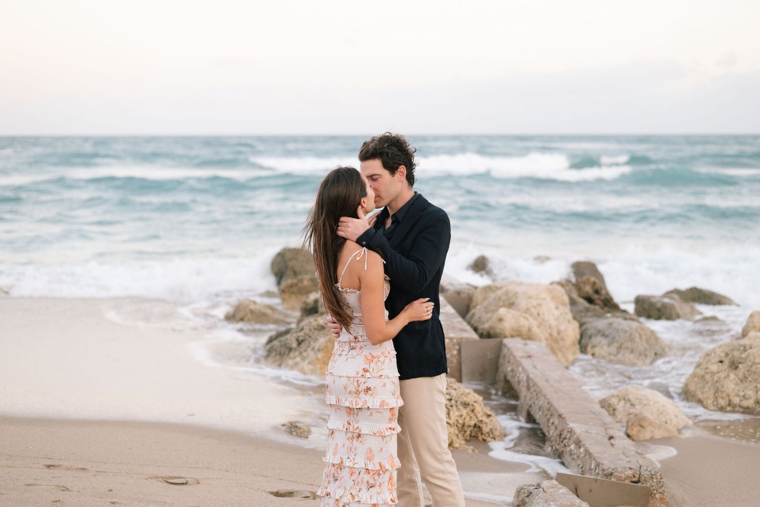 A couple shares a romantic moment on a beach with rocky outcrops and crashing waves in the background.