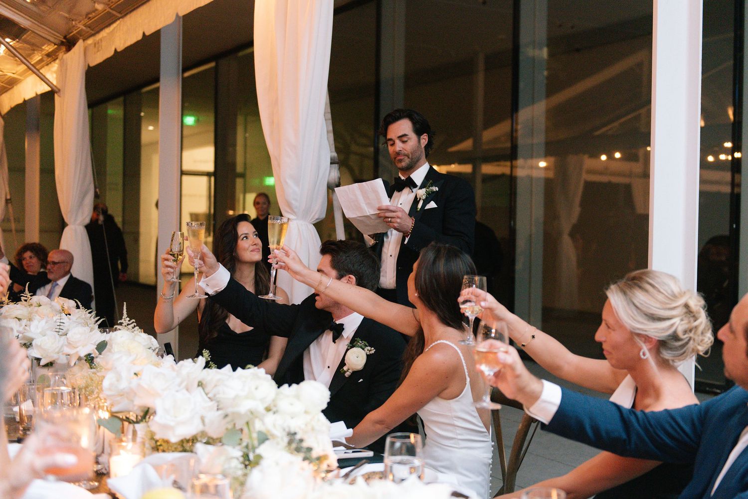 Wedding reception guests toast at elegant tables decorated with white floral centerpieces.