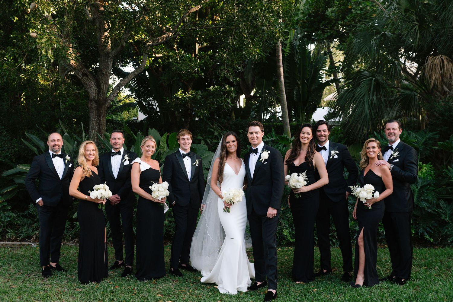 Large wedding party group photo with bridesmaids in black dresses and groomsmen in tuxedos outdoors.