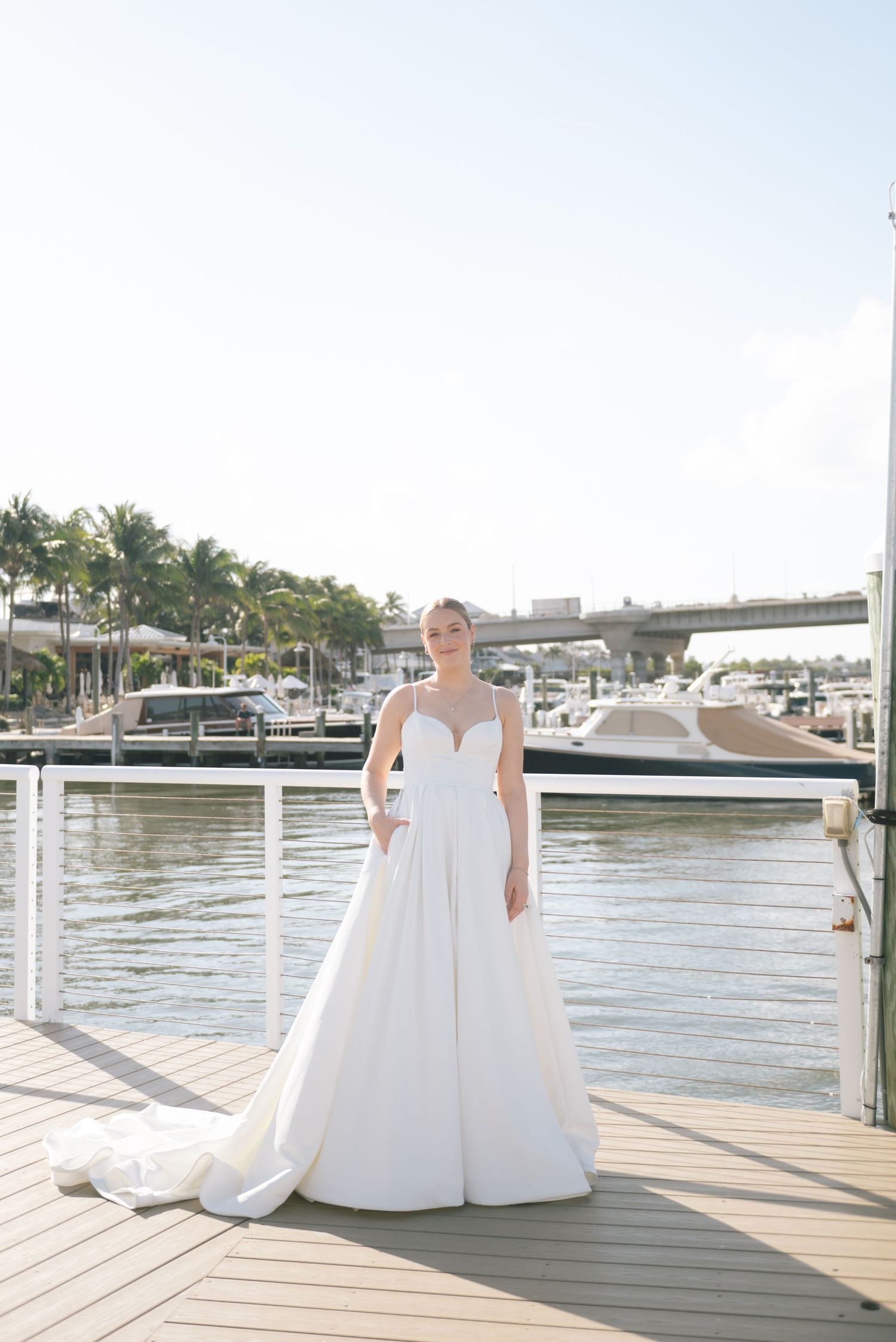 Two people in white dresses pose together on a waterfront dock with boats in the background.