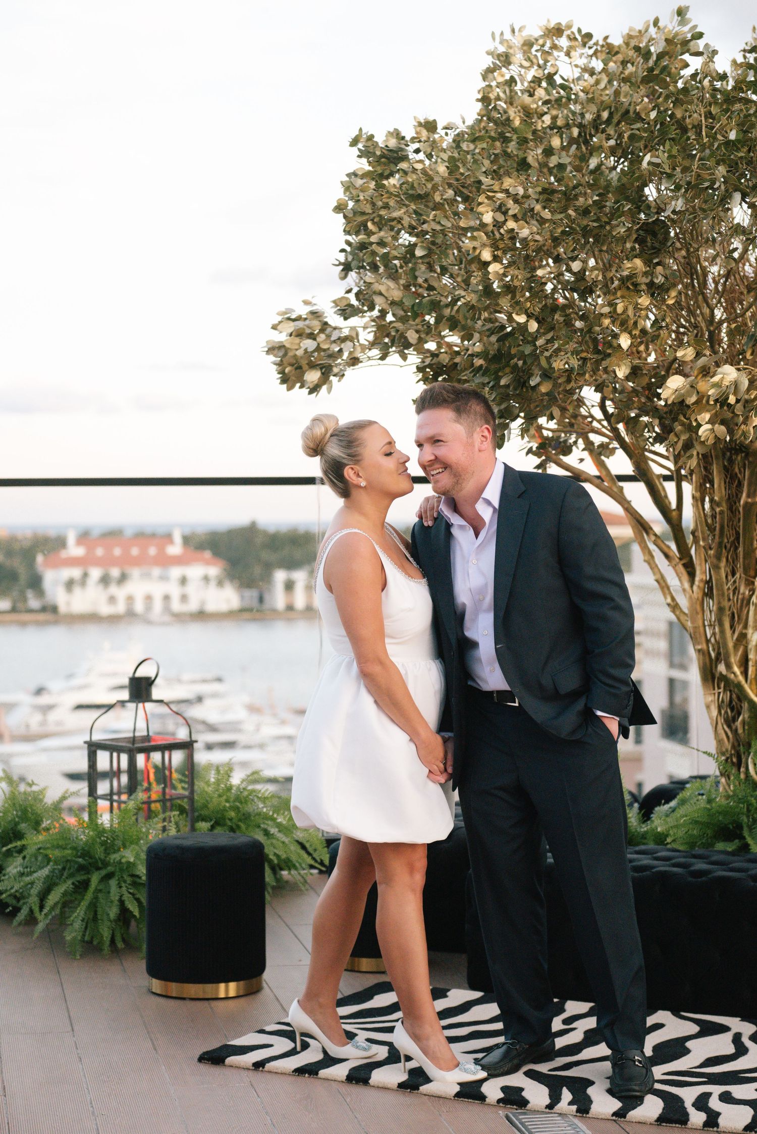 A couple shares a romantic moment on an outdoor terrace overlooking a marina with a decorative lantern nearby.