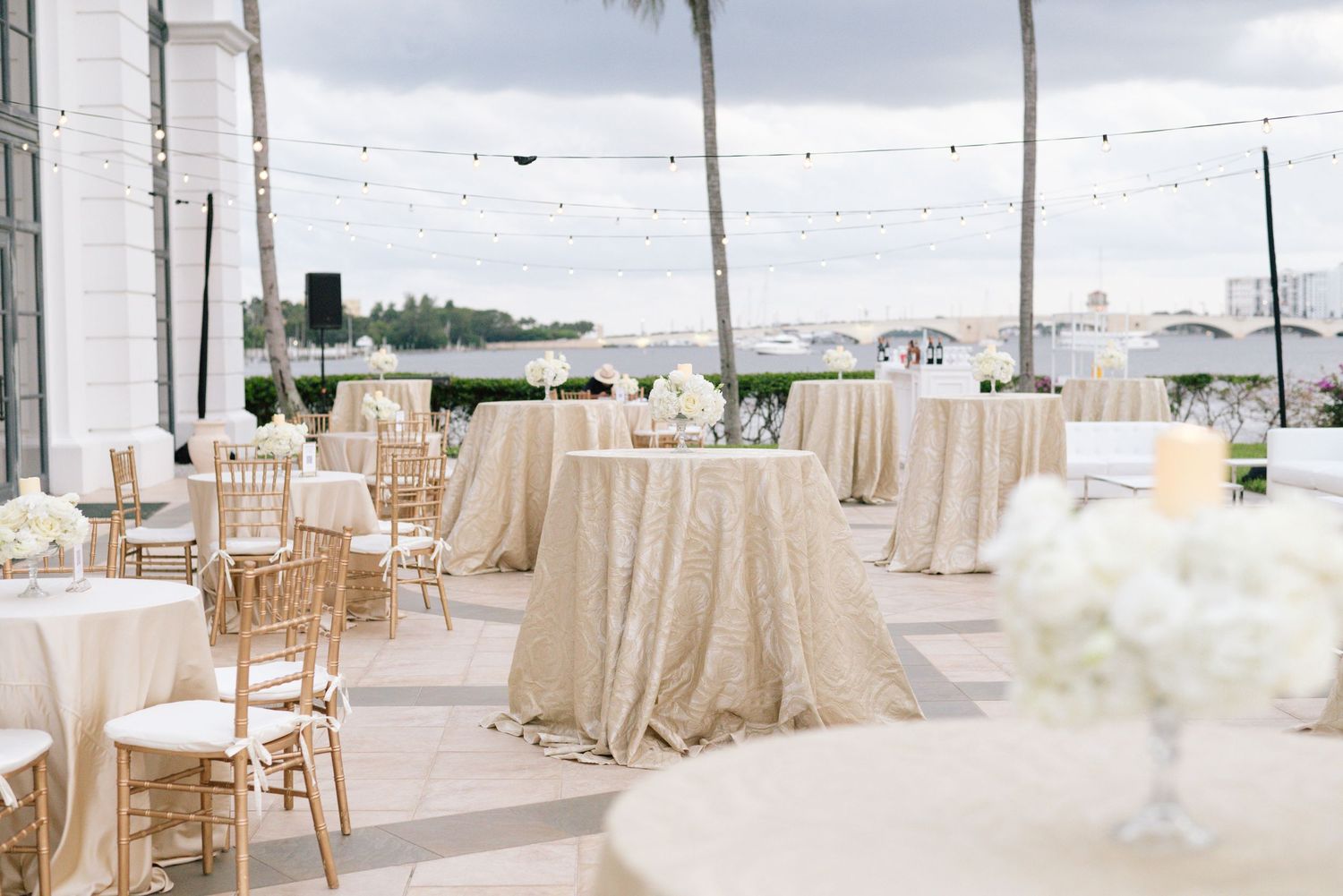 Outdoor wedding reception with gold sequin tablecloths, chiavari chairs, and string lights by waterfront.