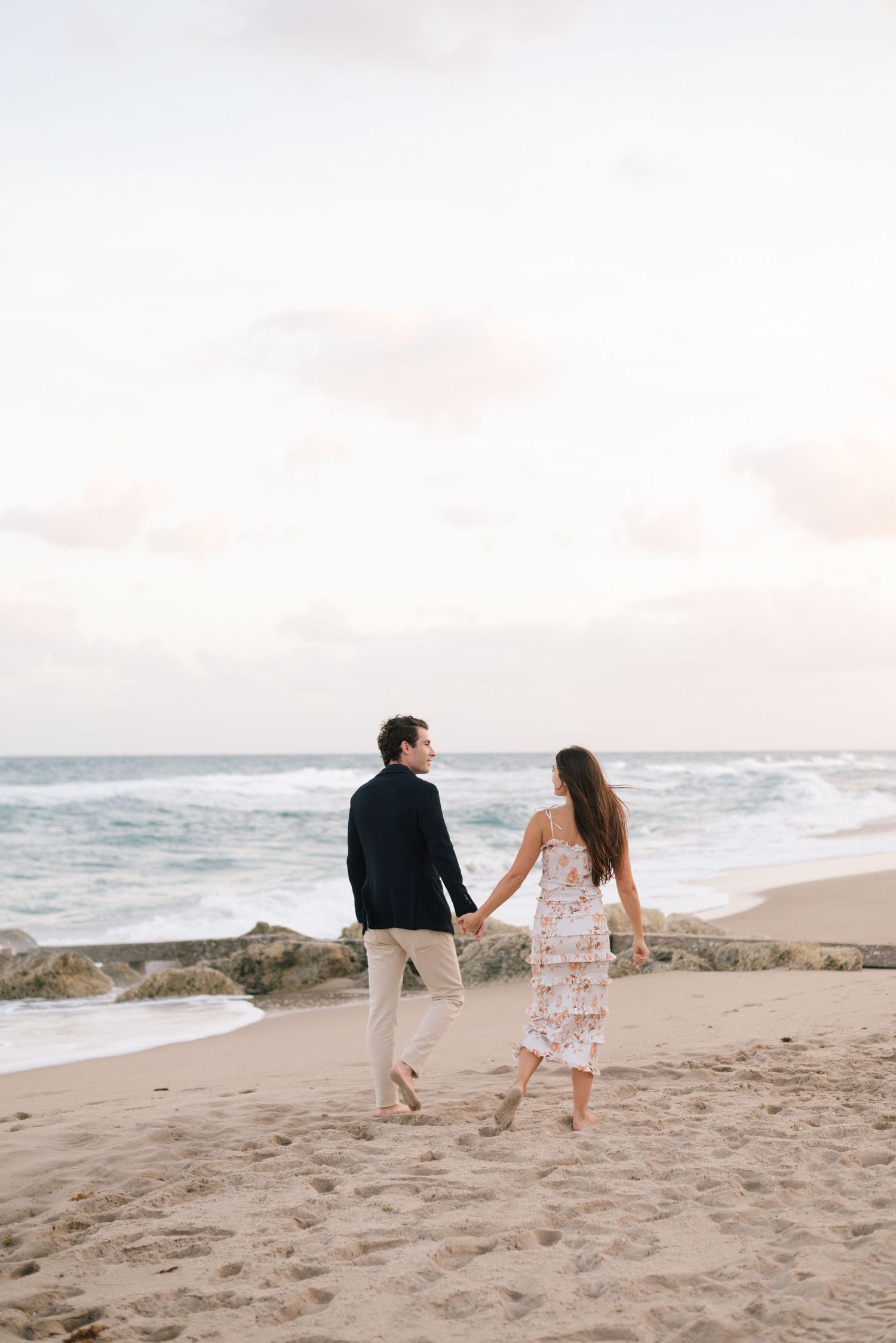 A couple strolls hand in hand on a sandy beach with gentle waves and cloudy skies in the background.