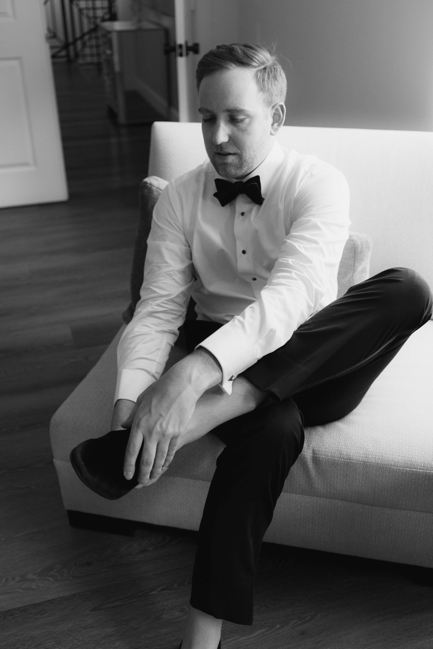 A person in formal attire sits on a white couch in a contemplative pose.
