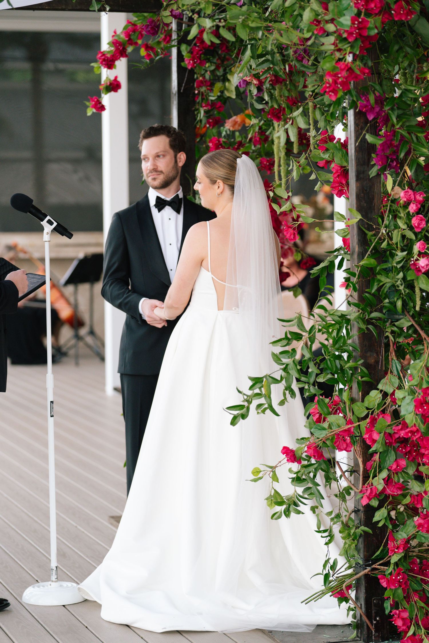 A wedding couple poses under an archway decorated with vibrant red climbing roses on a white porch.
