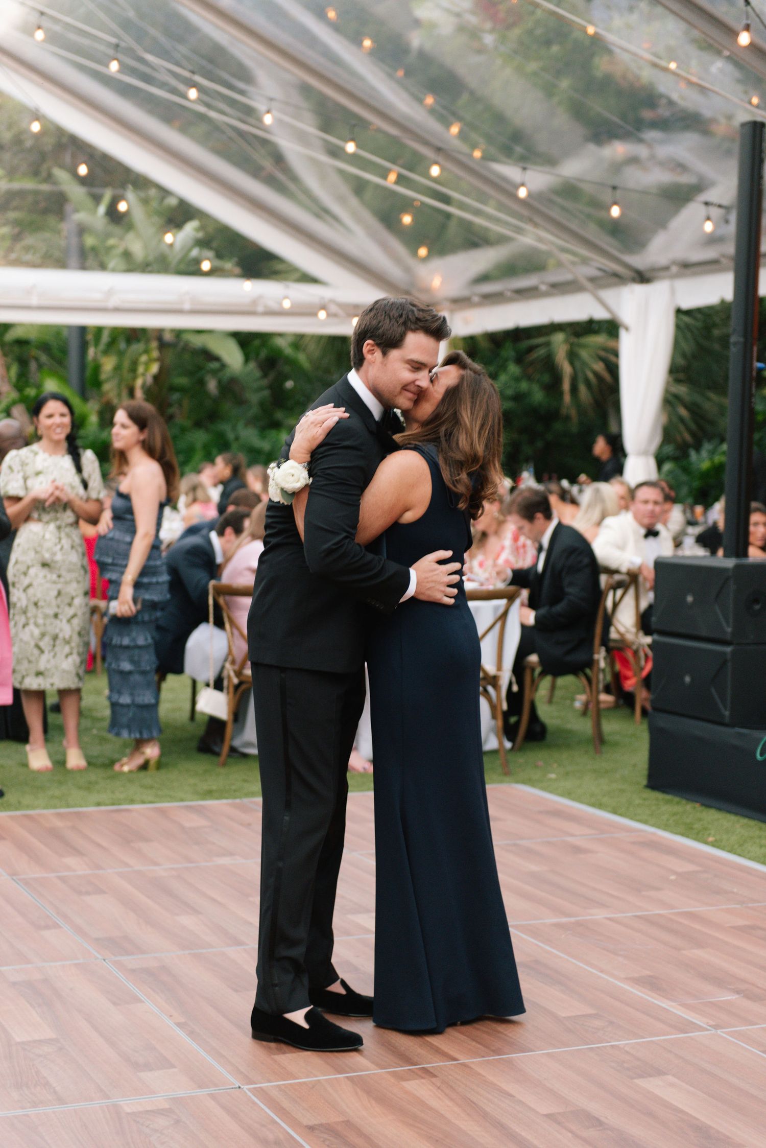 Couples dancing together on a wooden floor under a clear tent at an evening reception.