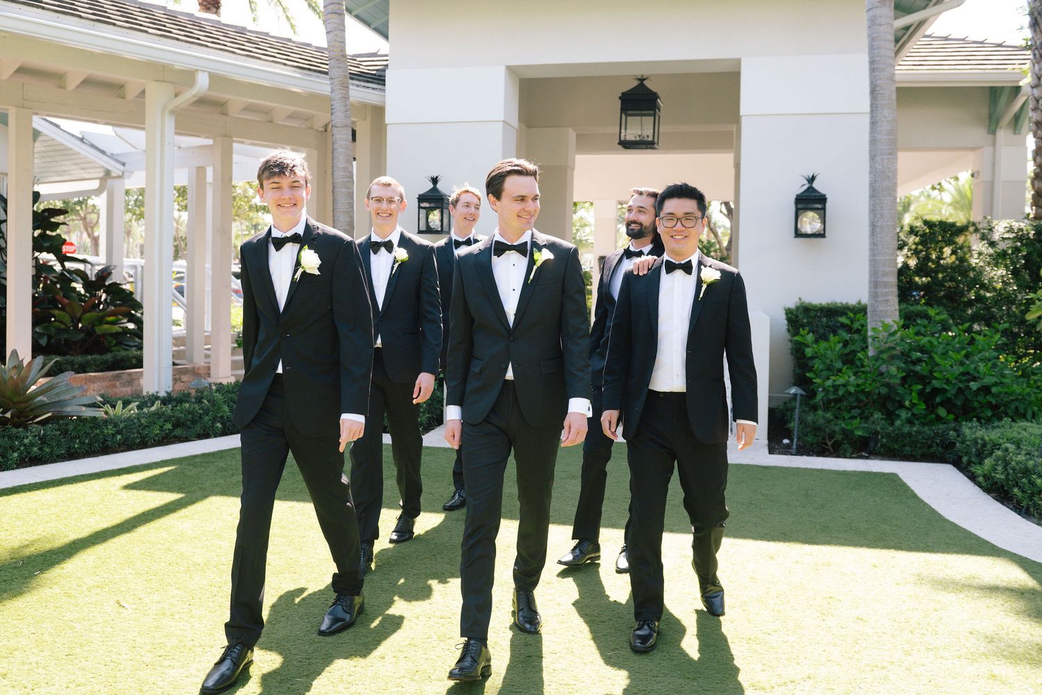 Groomsmen in black tuxedos walking along covered outdoor walkway.