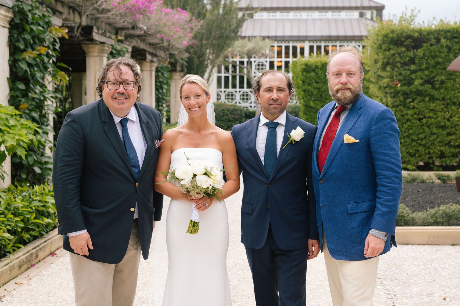 Small wedding group poses together on stone walkway with greenhouse and gardens in background.