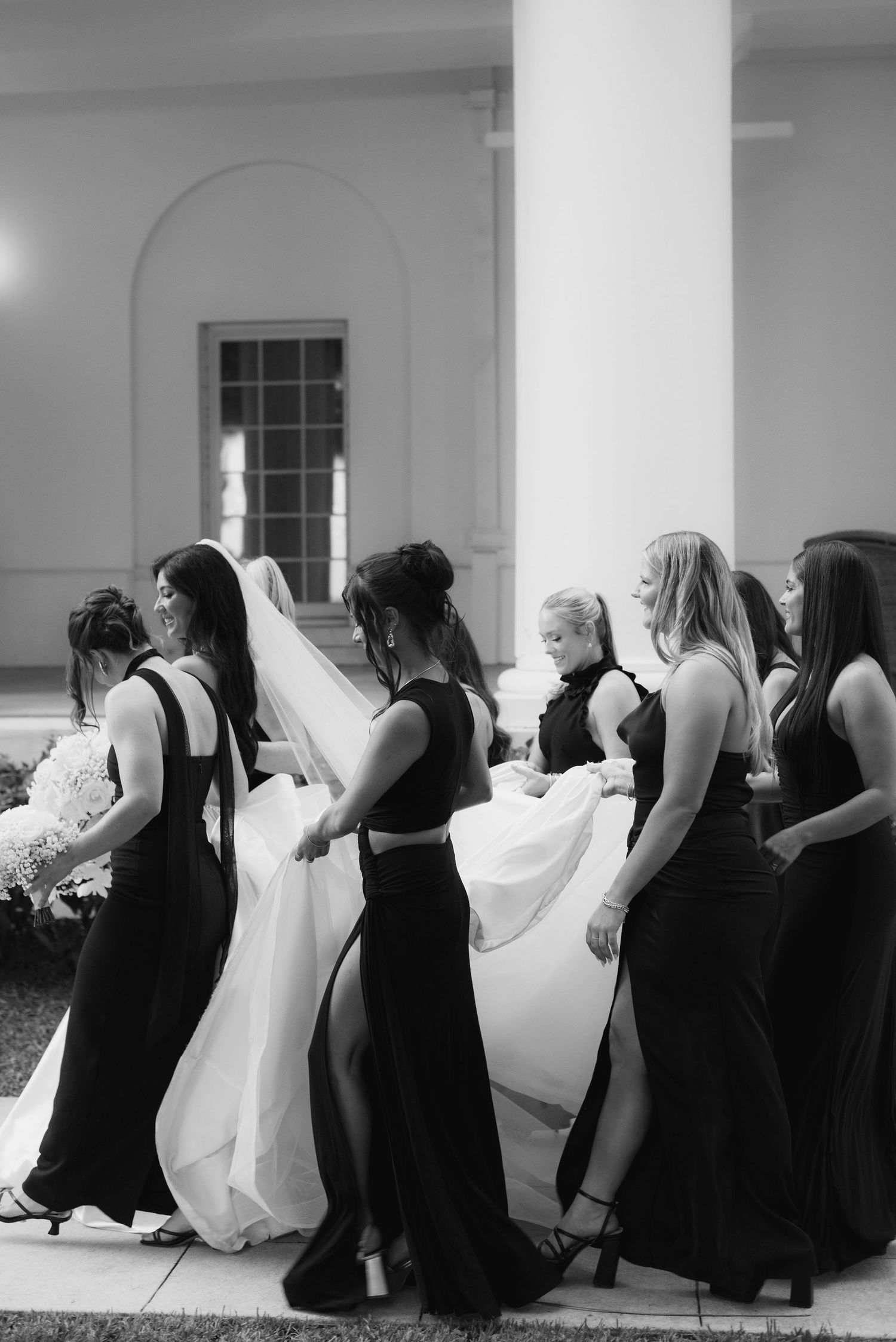 Bridesmaids in black dresses assist with a white wedding gown in front of columns.