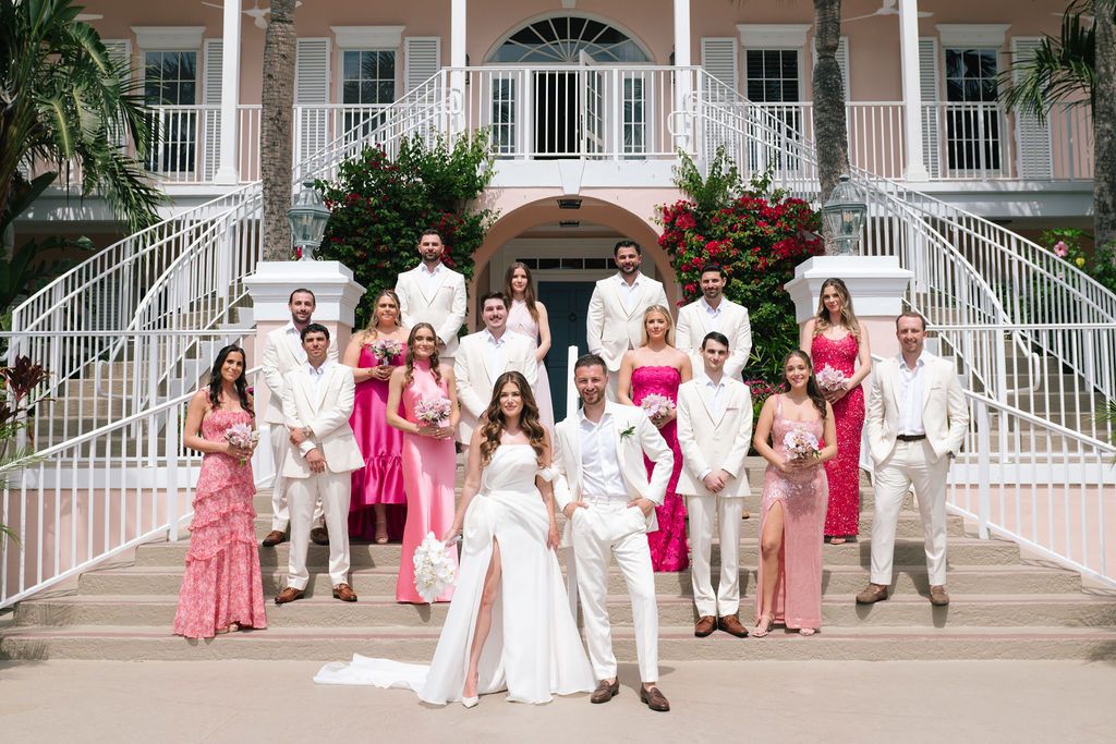 Large wedding party in pink dresses and white suits gathered on mansion steps for formal photo.