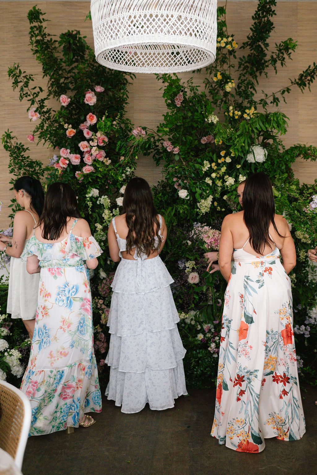 Bridesmaids in floral print dresses pose against a wall with climbing roses and white wicker pendant lights.