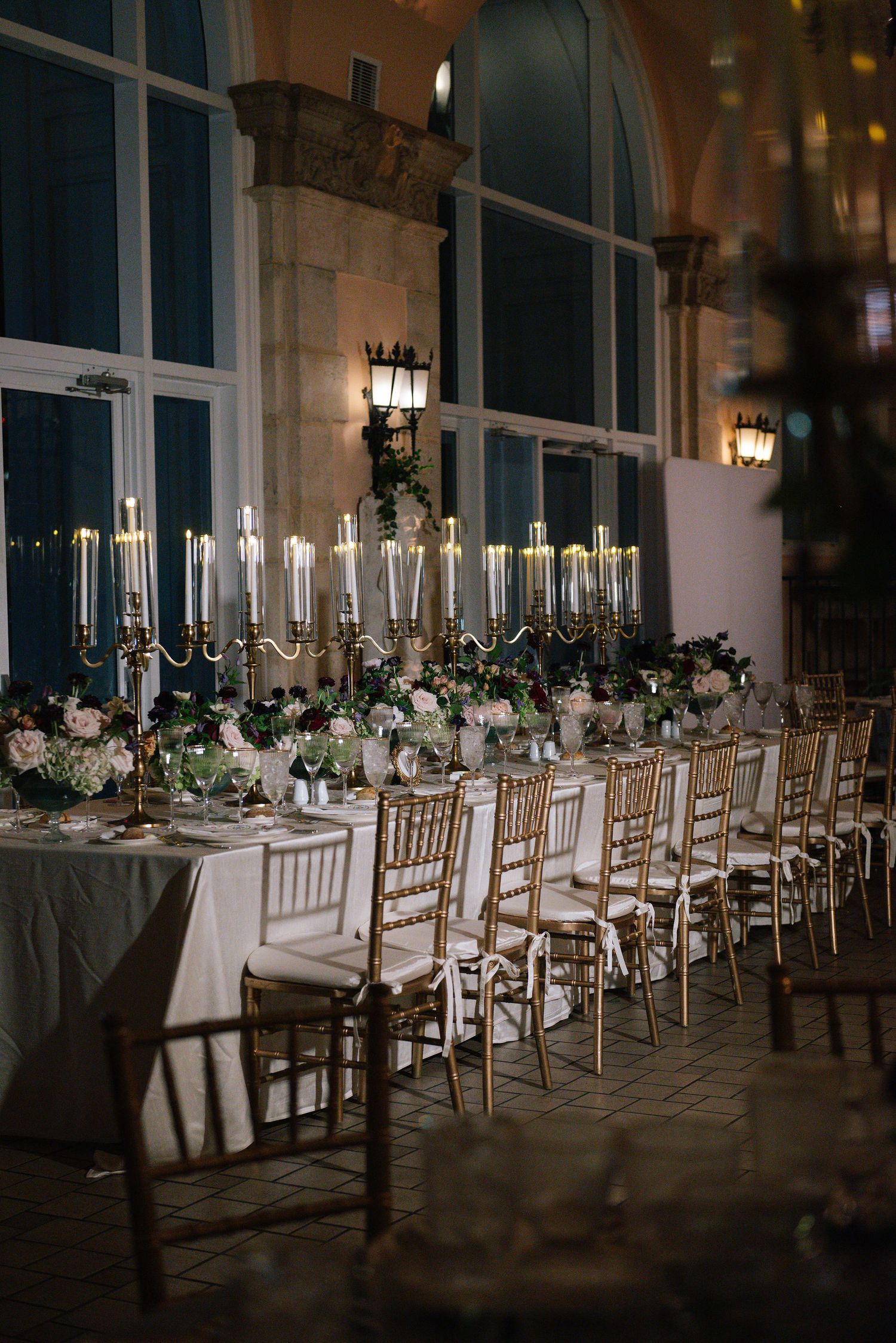 Dimly lit wedding reception tables with gold chairs and tall candelabras creating an intimate atmosphere.