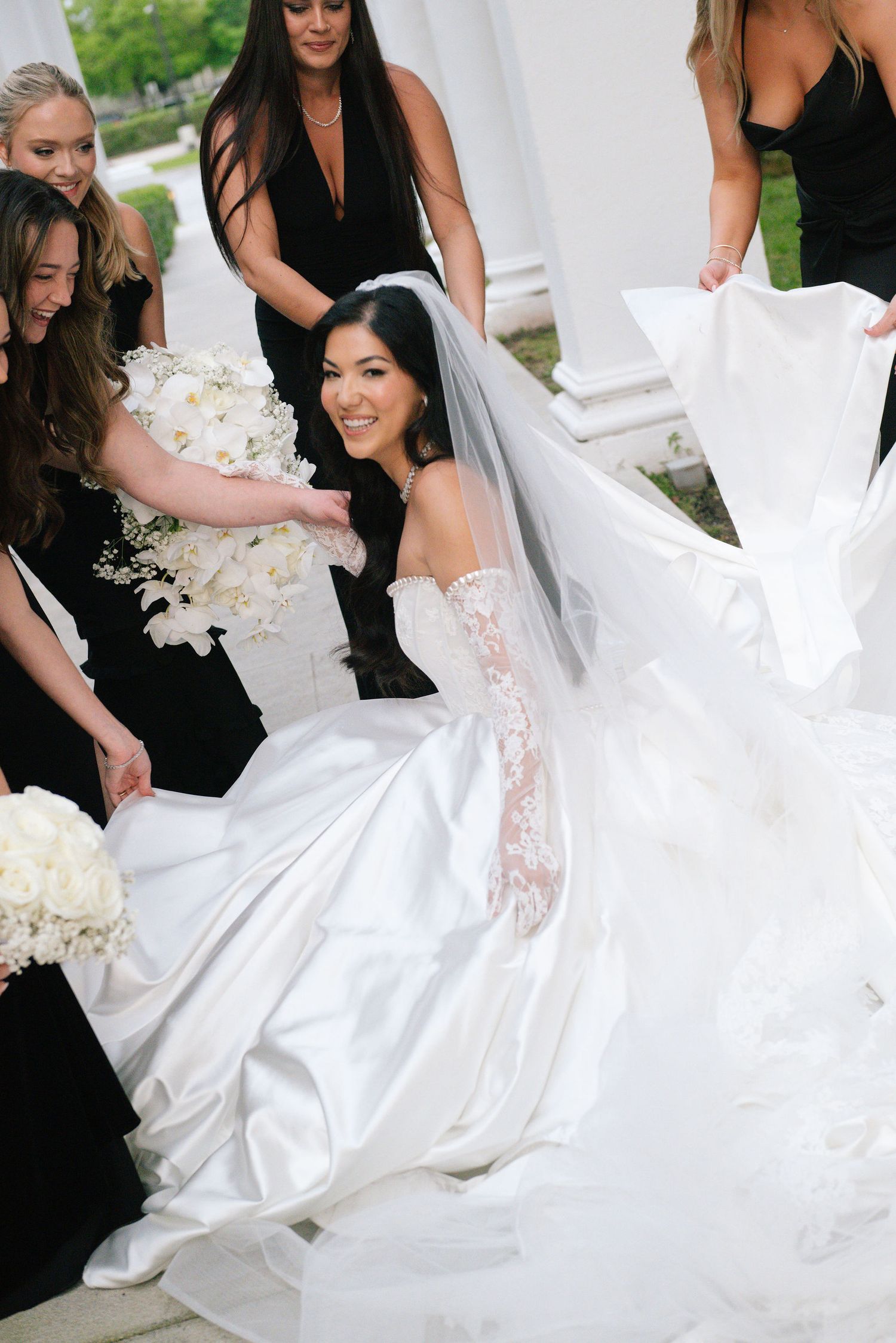 Bridesmaids in black dresses help adjust a flowing white wedding gown.