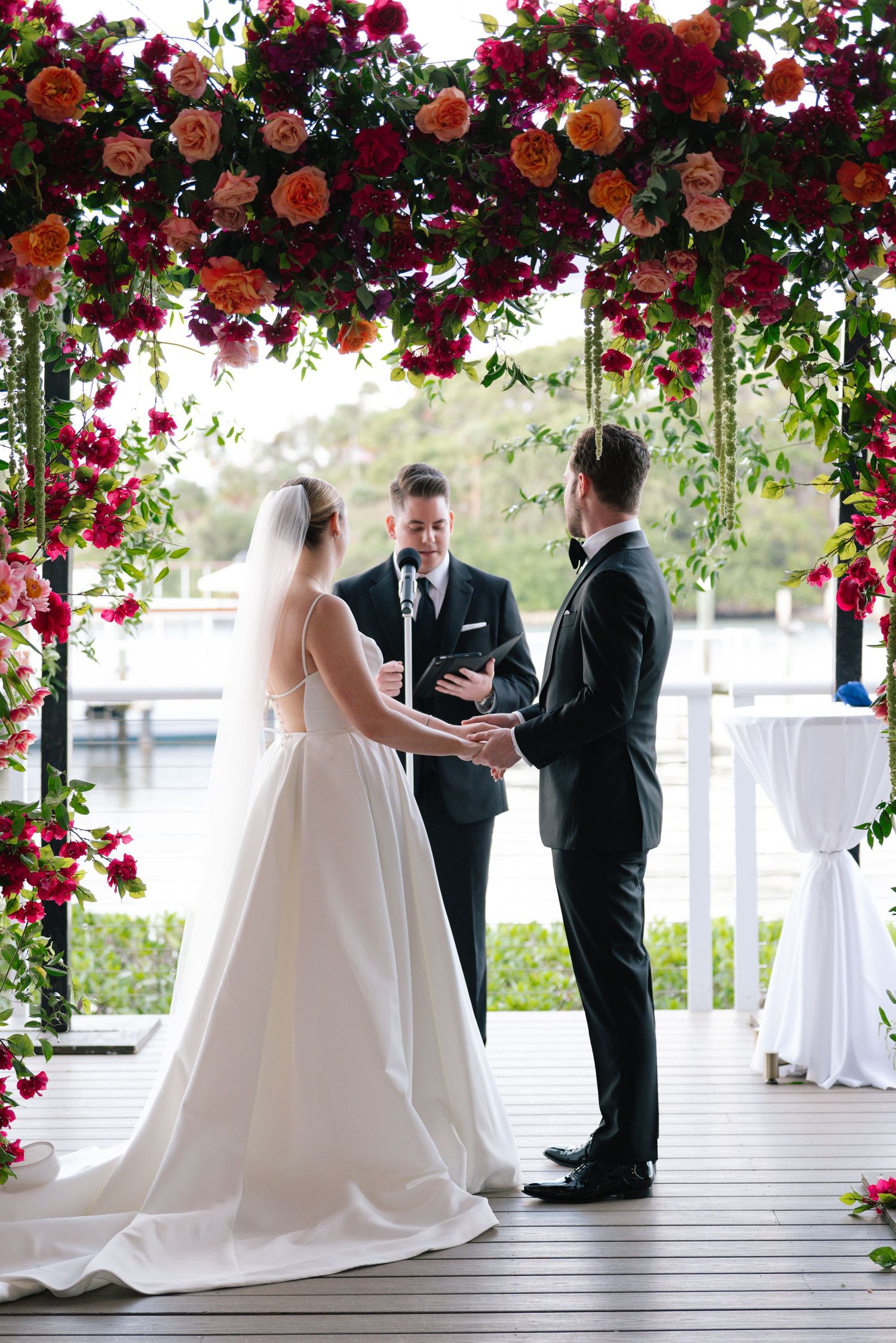 A wedding ceremony takes place under a beautiful archway decorated with vibrant red and pink roses.