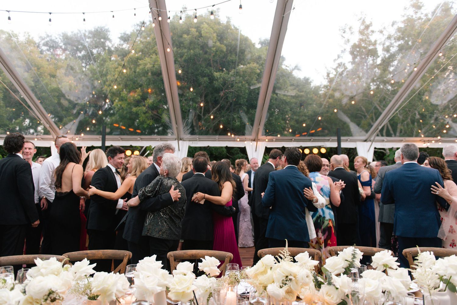 Wedding guests celebrate and dance together under an elegant white tent during an outdoor reception celebration.