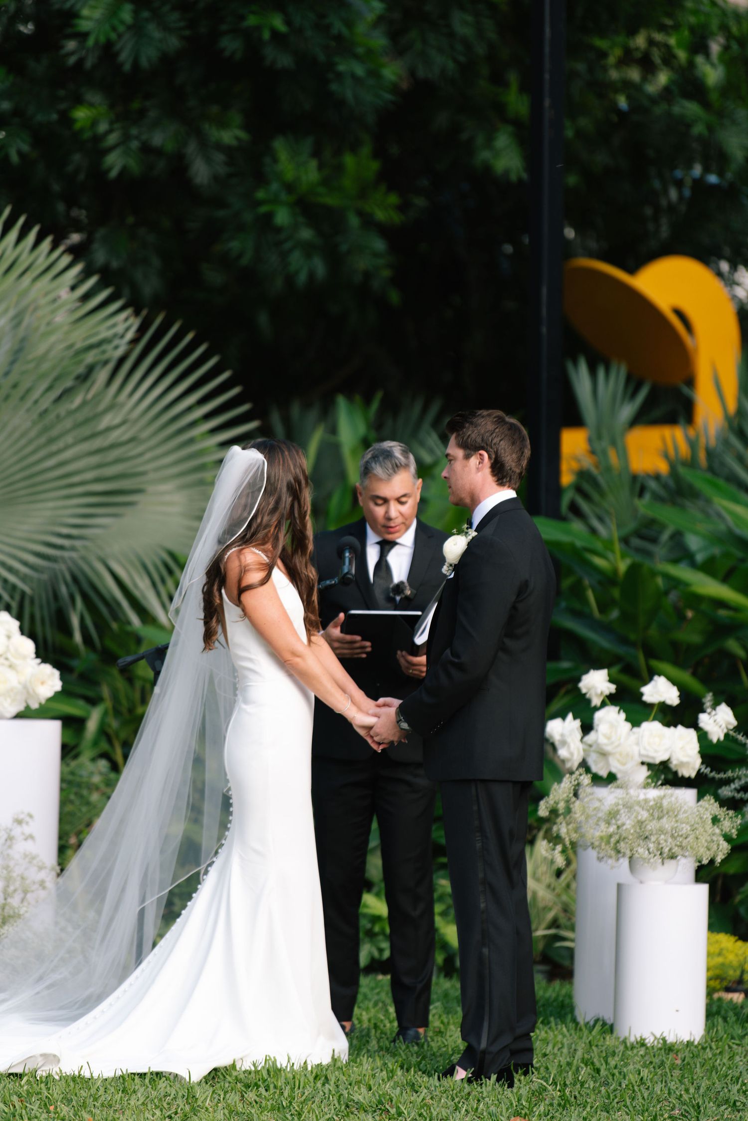 A wedding ceremony unfolds on a grassy lawn with tropical plants and white floral arrangements.
