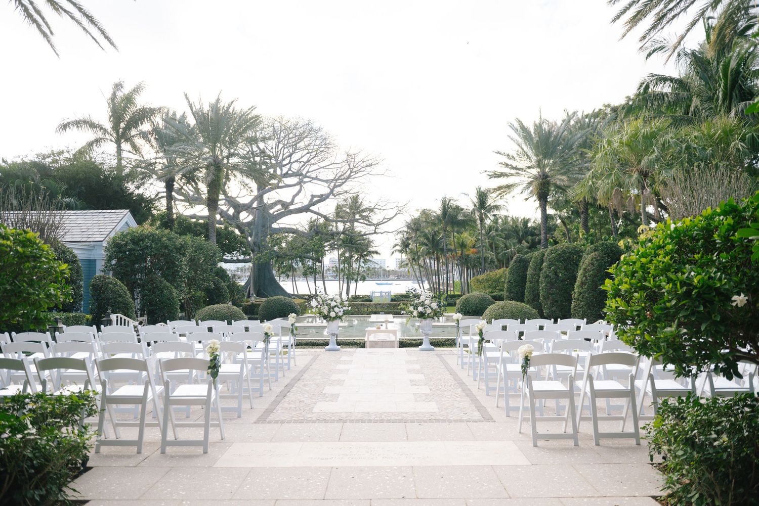 Elegant outdoor wedding ceremony setup with white chairs and tropical palm trees leading to oceanfront view.