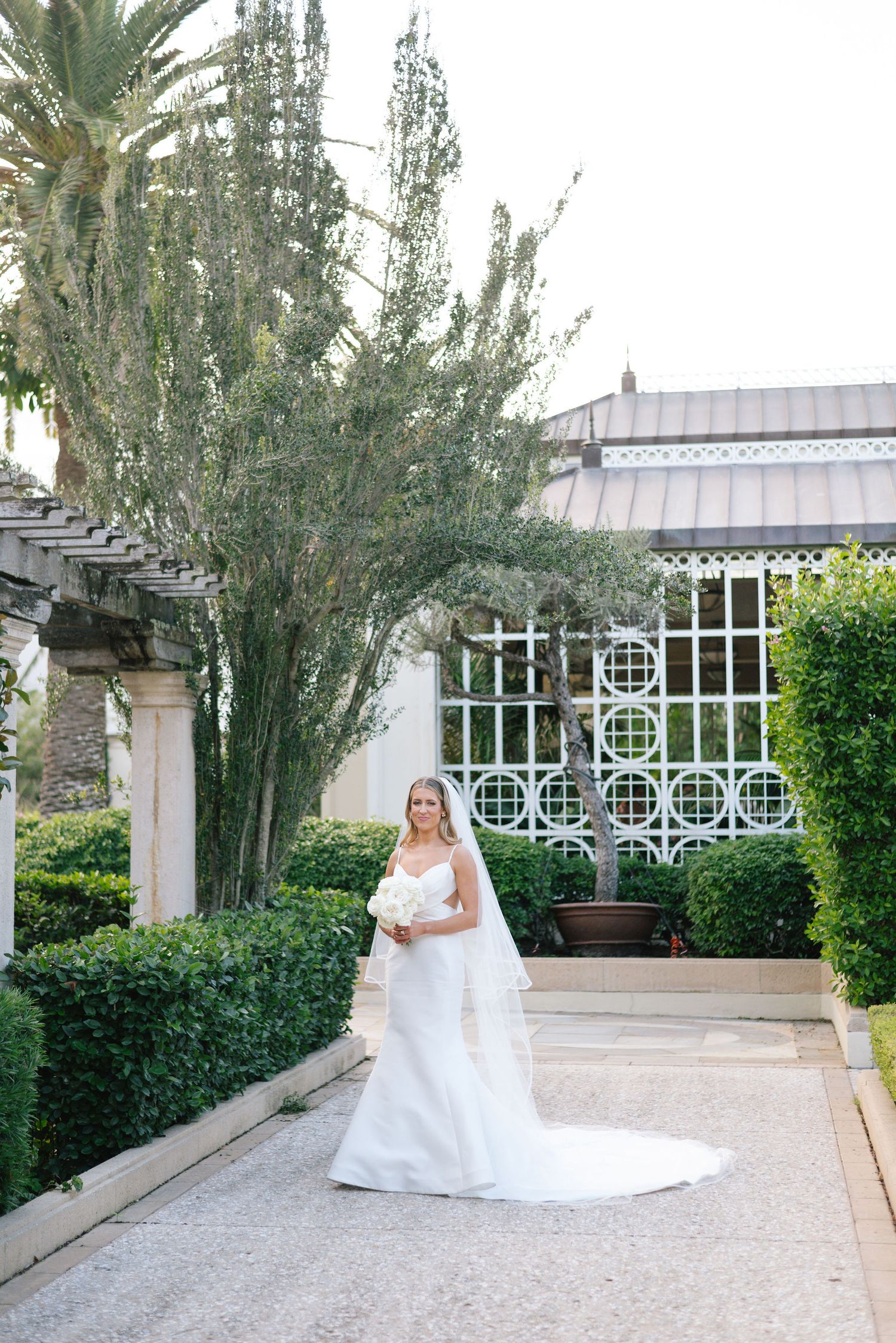 A bride in a white wedding gown poses in a garden courtyard with ornate architecture and palm trees.
