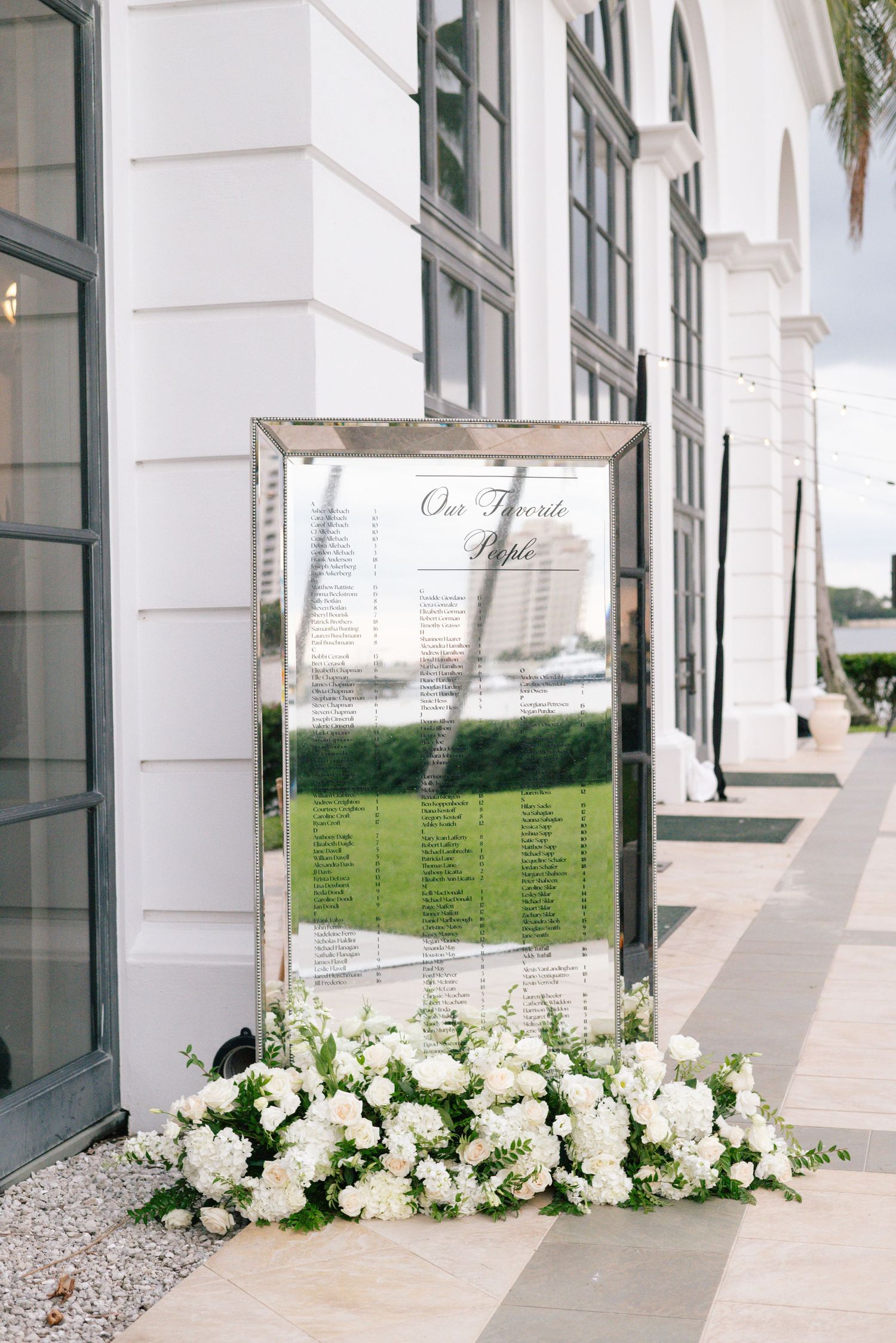 Mirrored seating chart sign surrounded by white floral arrangement at outdoor wedding venue.