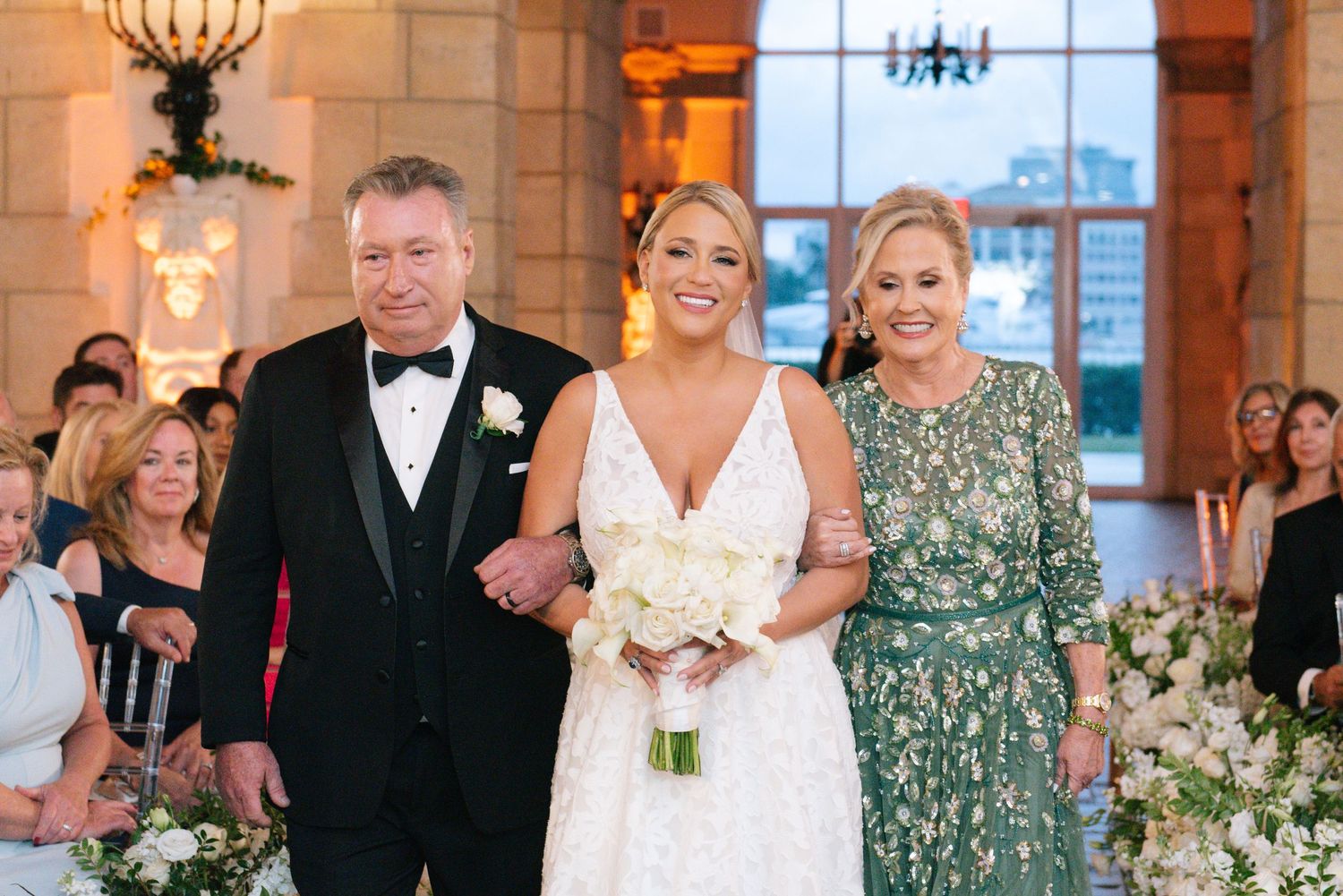 Bride in white gown walks down aisle with parents during elegant wedding ceremony with floral decorations.