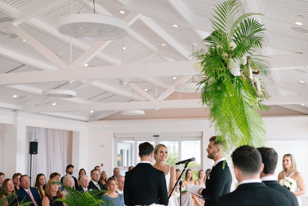 Panoramic view of wedding ceremony in modern white venue with tropical greenery.